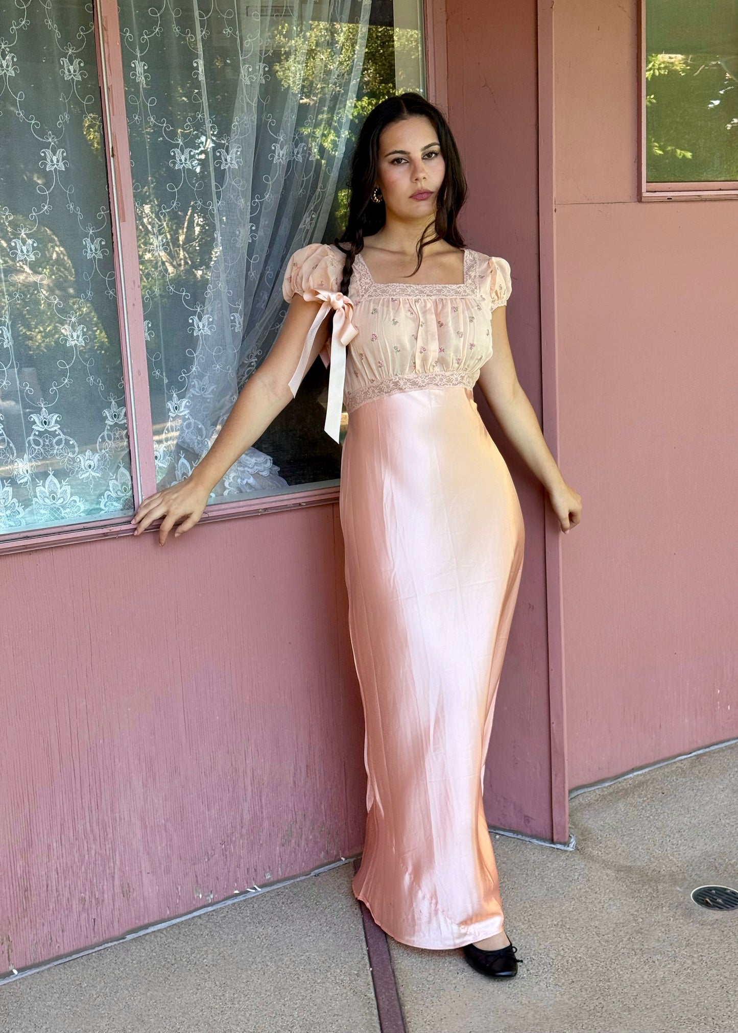 Woman in a long, light-colored dress standing in front of a pink wall with a window.