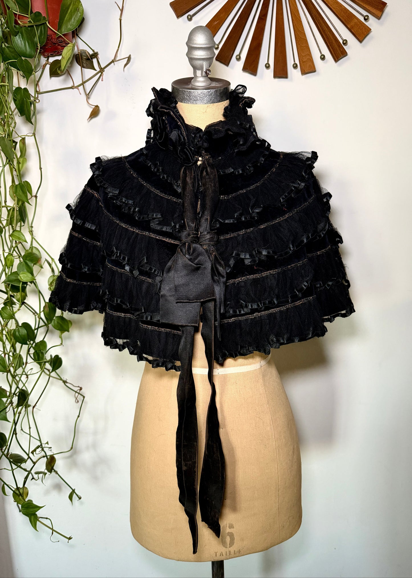 Black ruffled cape on a mannequin with a decorative wall and plant in the background