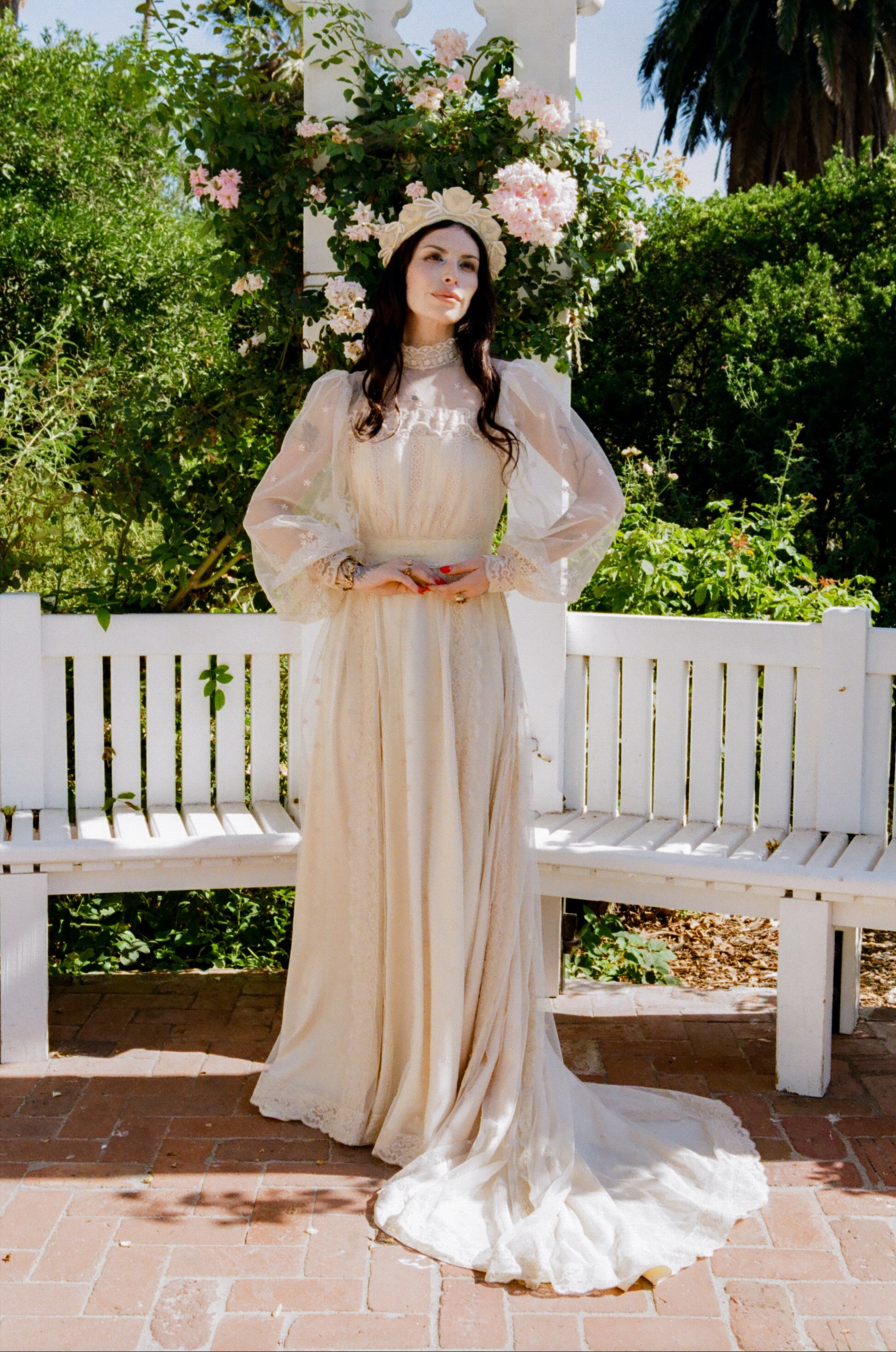 Woman in a vintage-style dress with floral headpiece standing in a garden setting.