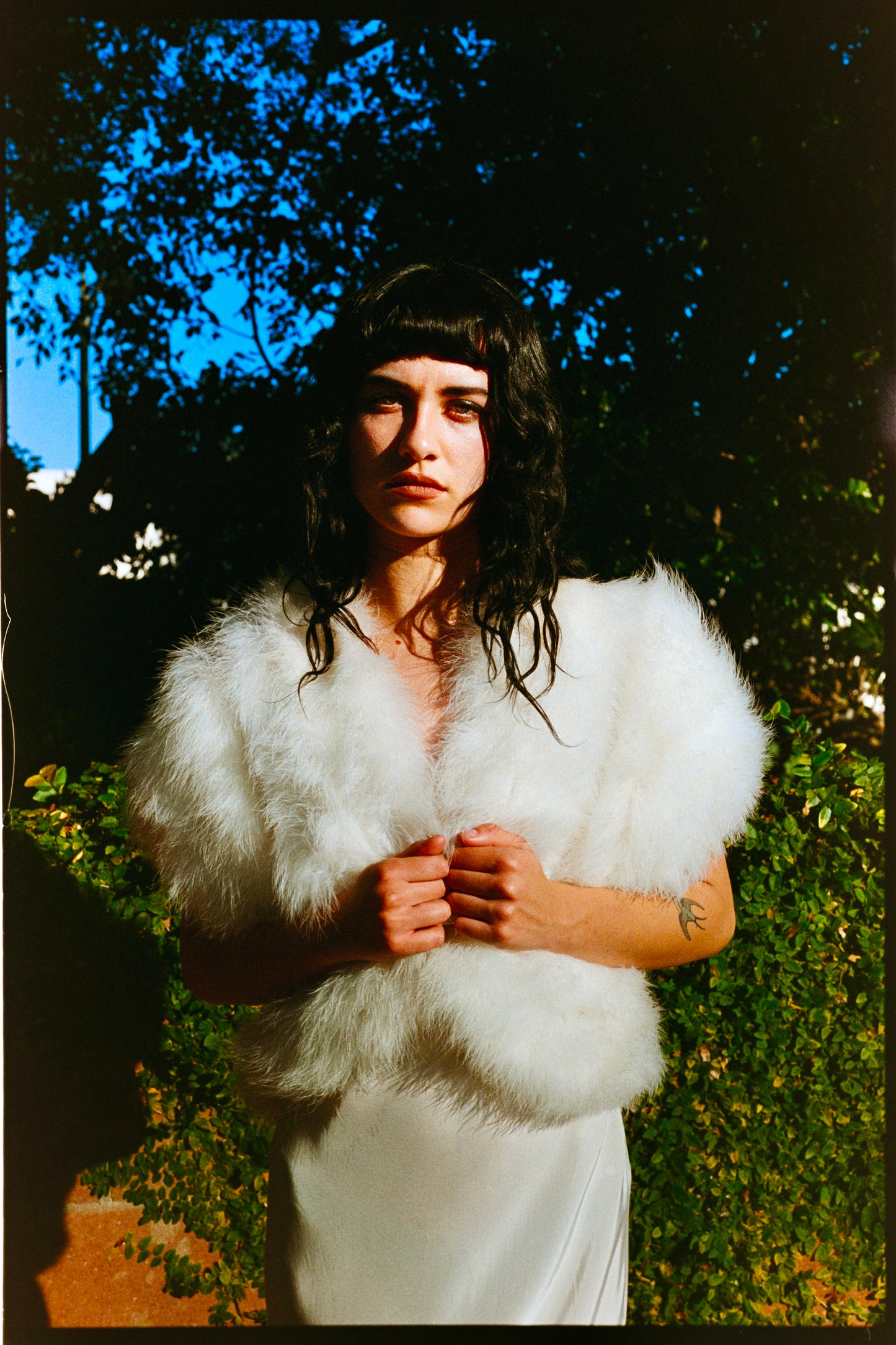 Person wearing a white marabou vest outdoors with trees and a wooden fence in the background