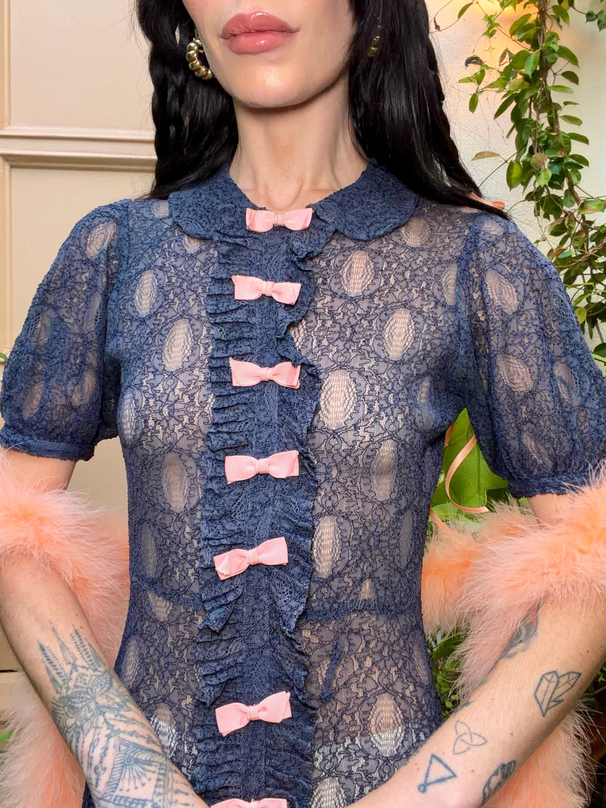 A woman wearing an antique 1930's navy blue lace dress with baby pink bows placed from the neckline all the way to the bottom of the dress, right in the center of the dress. Romantic puff sleeves, bias cut silhouette. 