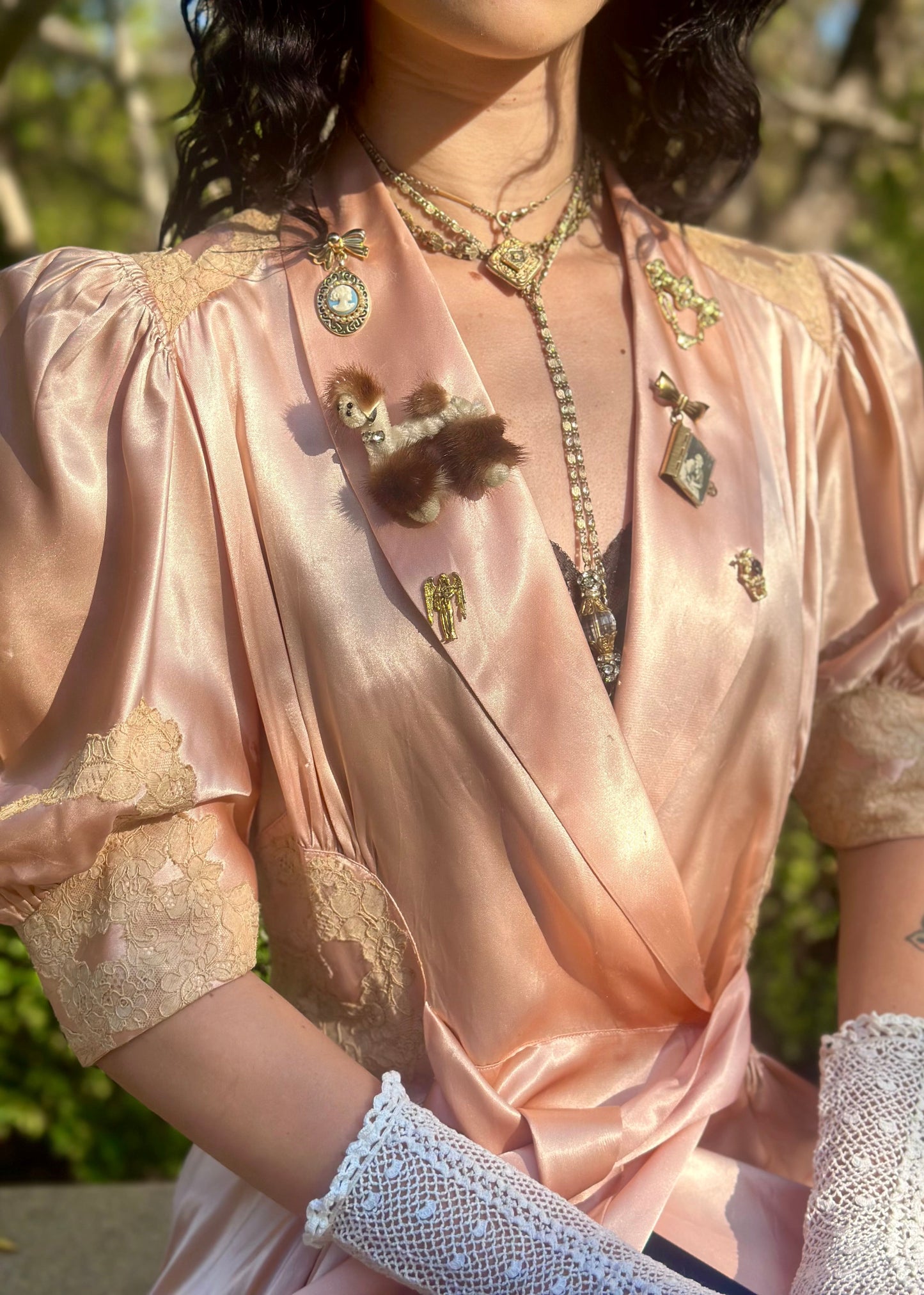 Woman in a 1940's dressing gown in peach pink with antique brooches standing between two metal arches in a garden.