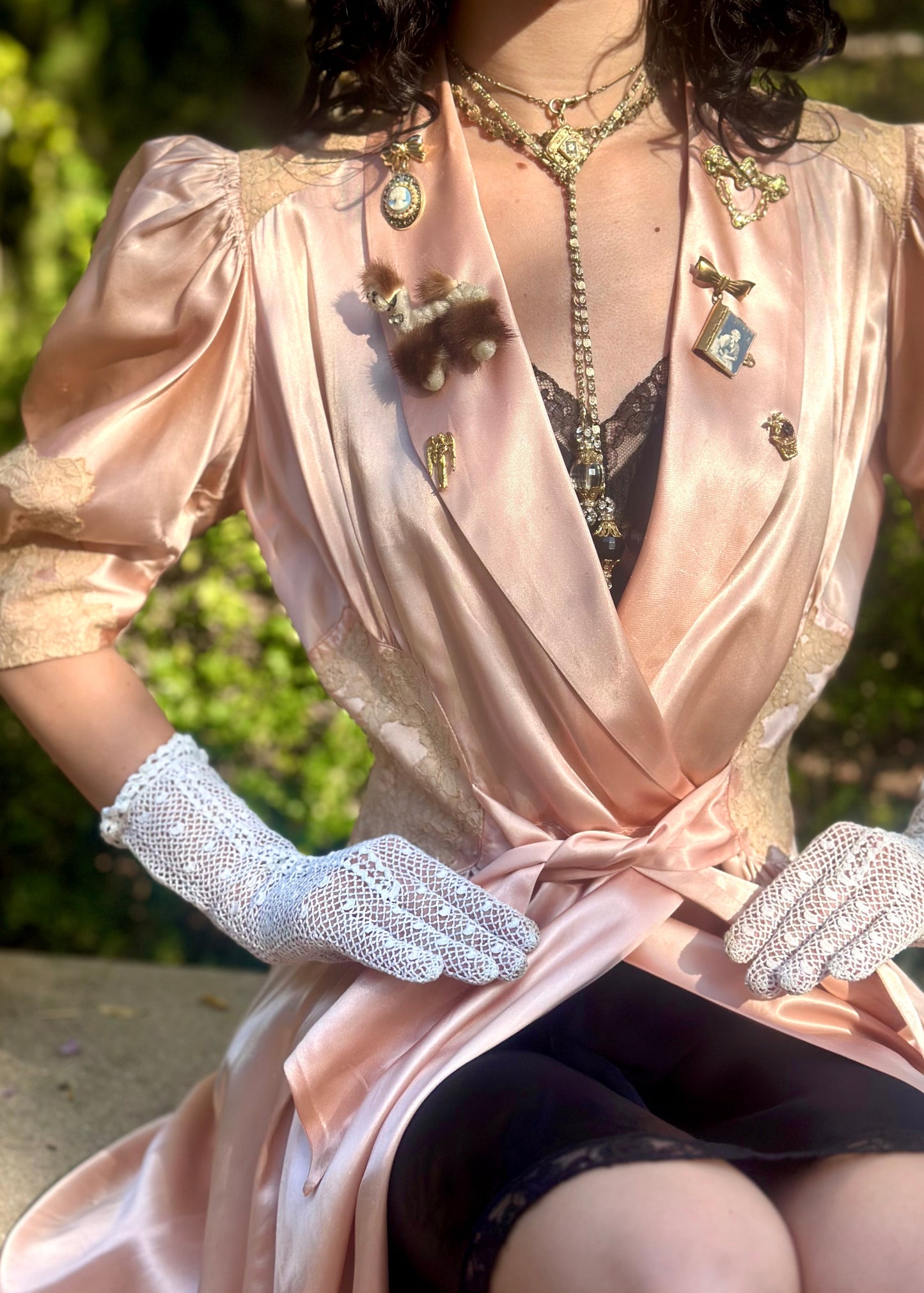 Woman in a 1940's dressing gown in peach pink with antique brooches standing between two metal arches in a garden.