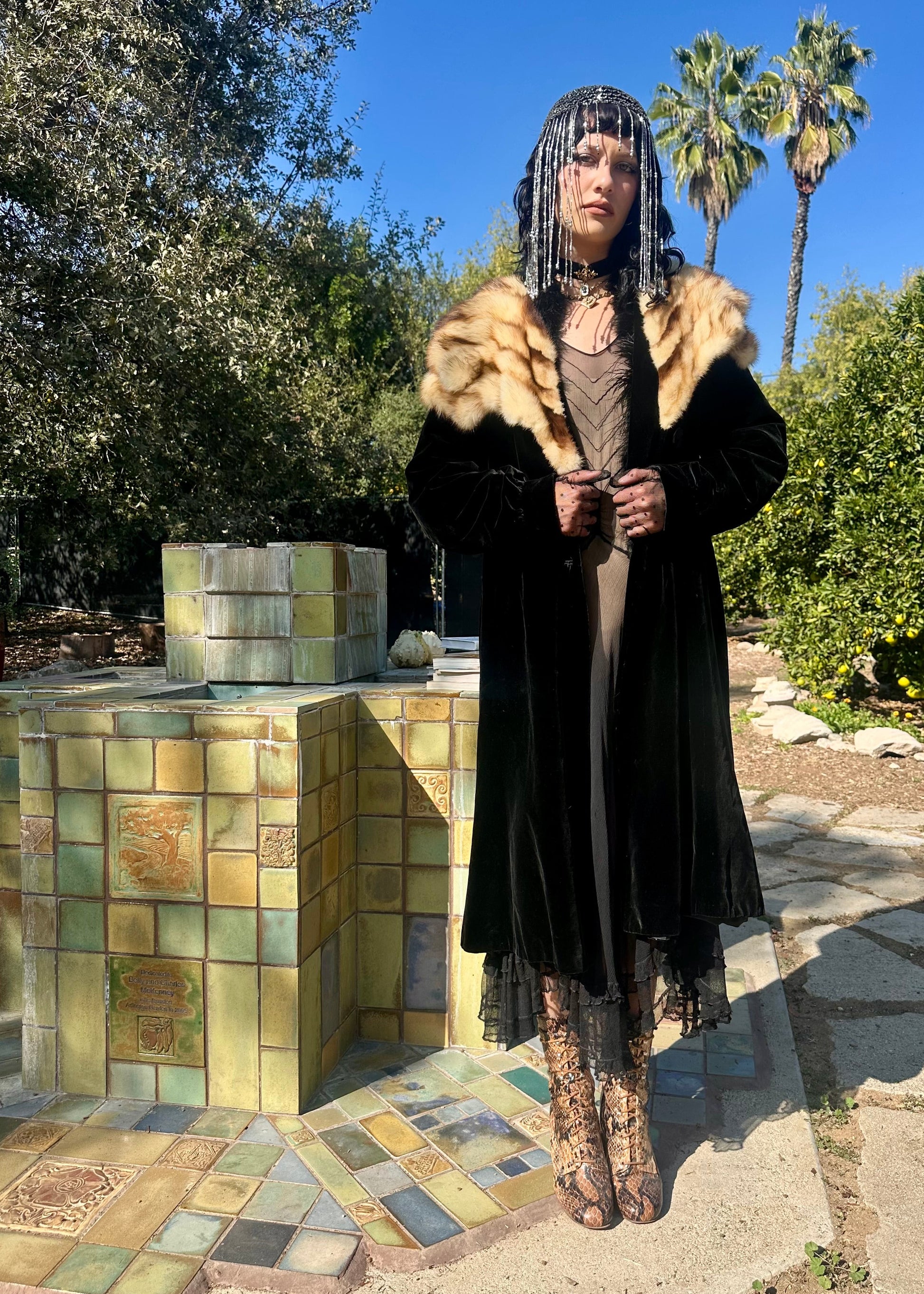 Person in an antique 1930's long black coat with fur trim standing next to a colorful tiled structure in a garden setting.