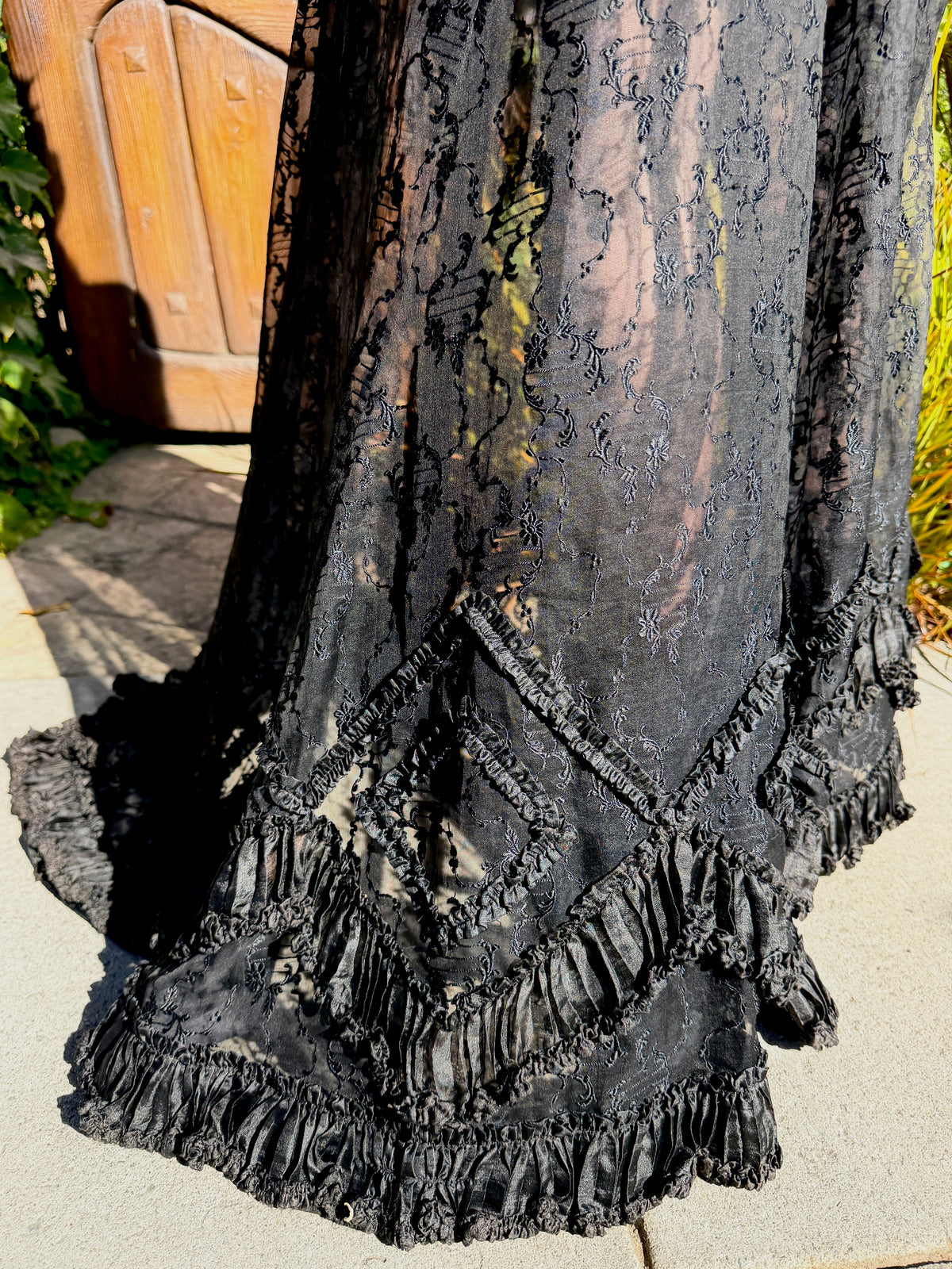 A close-up of a black lace and tulle dress skirt with tiered ruffles, photographed outdoors on a stone surface near a wooden door.