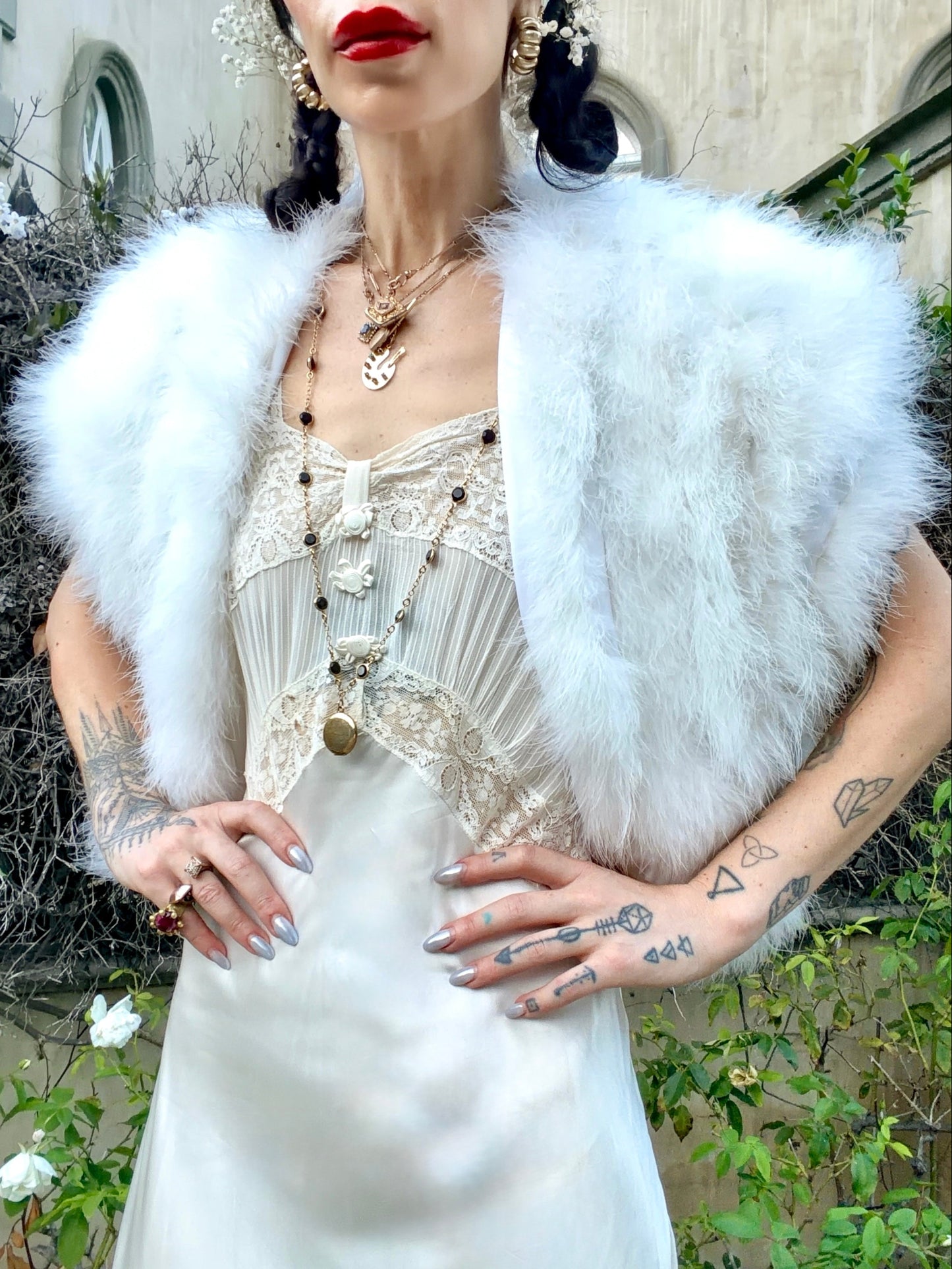 A woman wearing a fluffy white faux-fur shrug over a cream lace-trimmed dress, photographed outdoors with garden foliage and a building wall in the background.