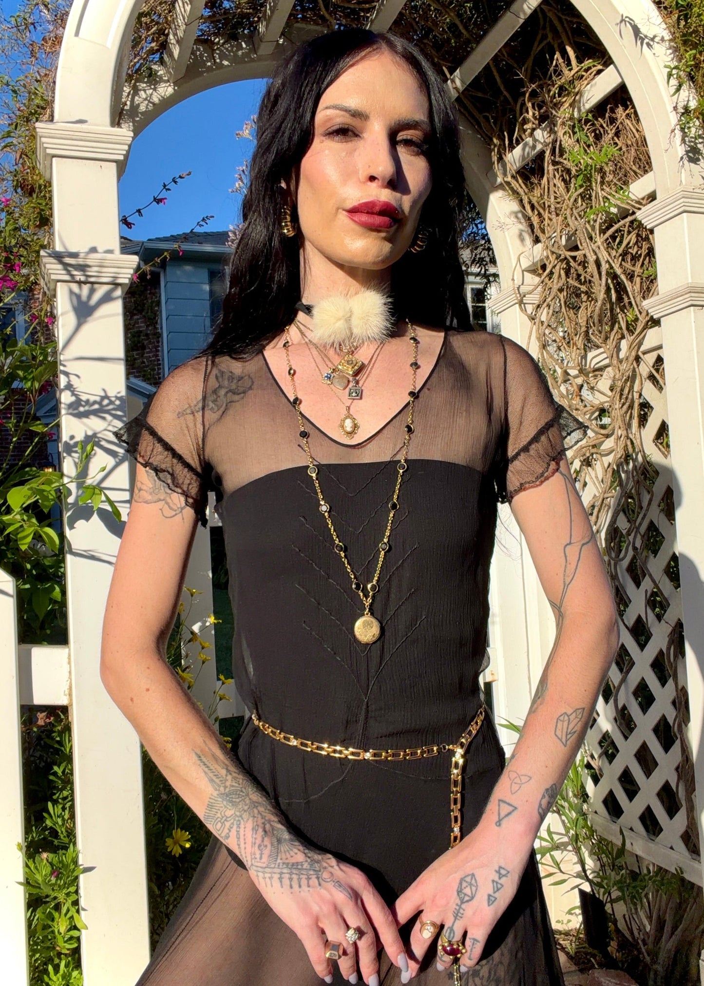 A woman stands outdoors under a white arched garden gate wearing a sheer black antique 1920's dress with a long black mesh skirt, layered gold necklaces, a gold chain belt, and beige high heels.