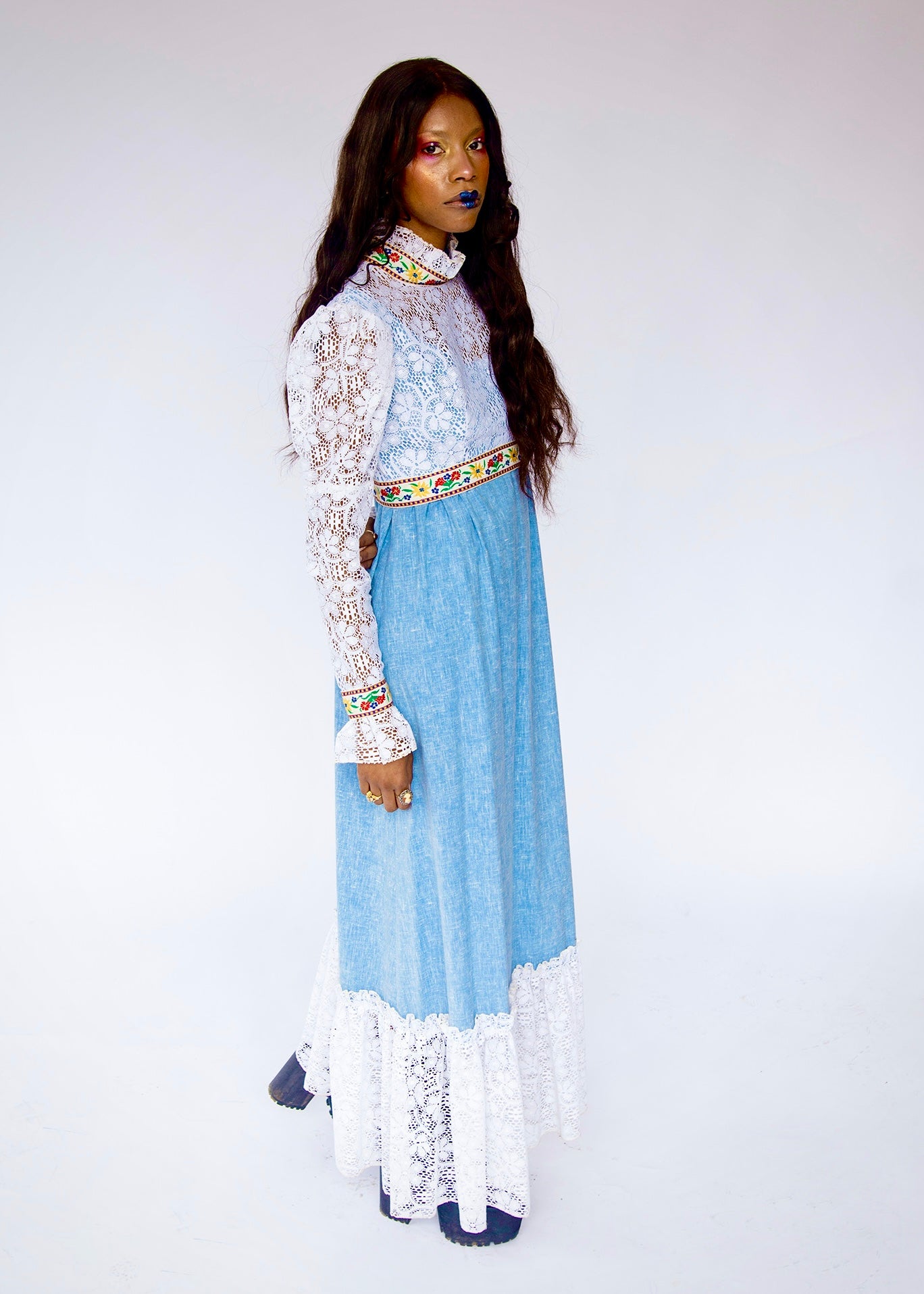 A model wearing a vintage 1970's prairie style boho hippie maxi dress made from denim and lace with mutton style long sleeve. Light blue denim maxi dress with intricate white lace overlays on the bodice and sleeves, a colorful patterned waistband, and a white lace hem, photographed against a plain white studio background.