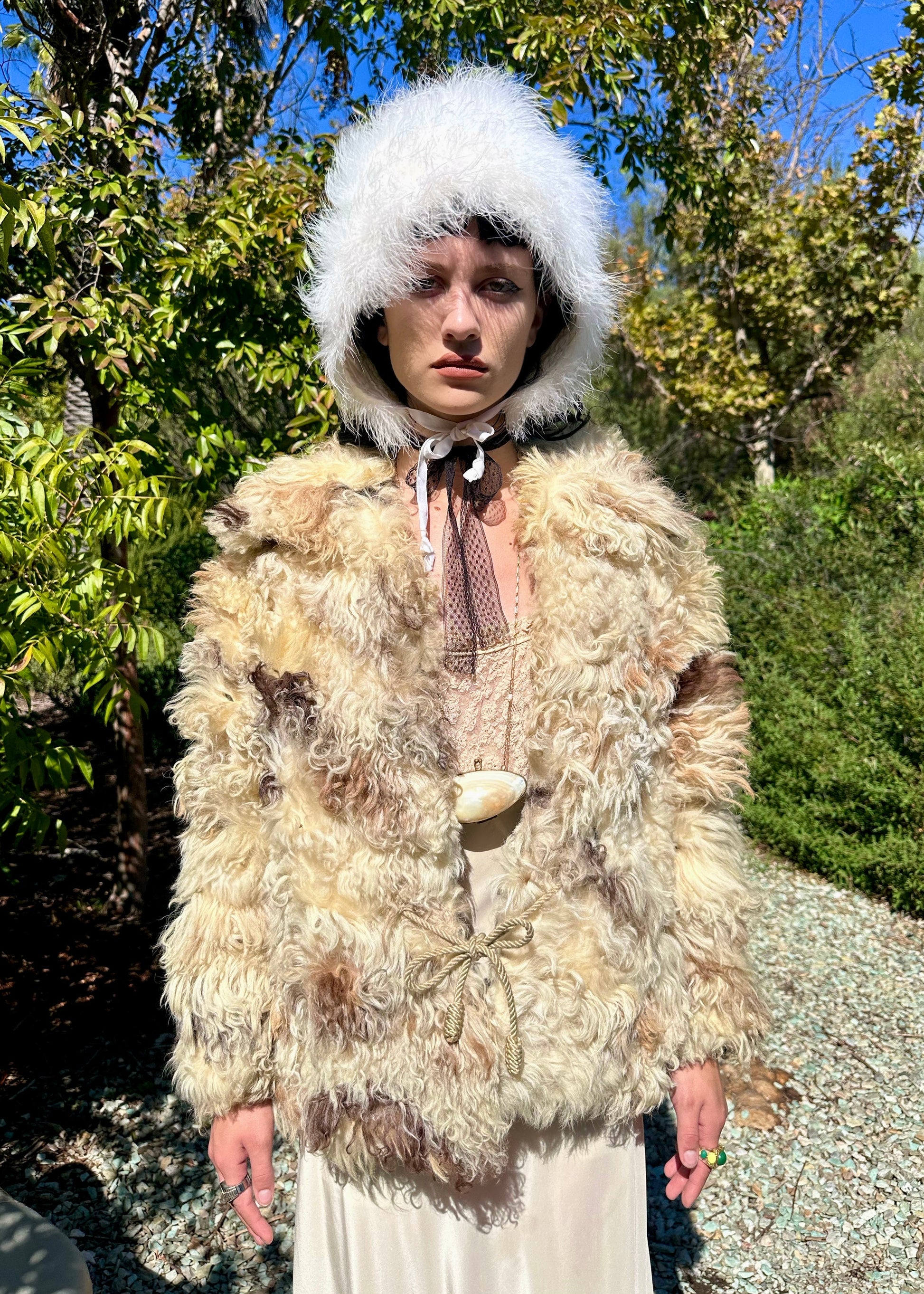 Person wearing a textured fur coat and white hood outdoors with greenery in the background