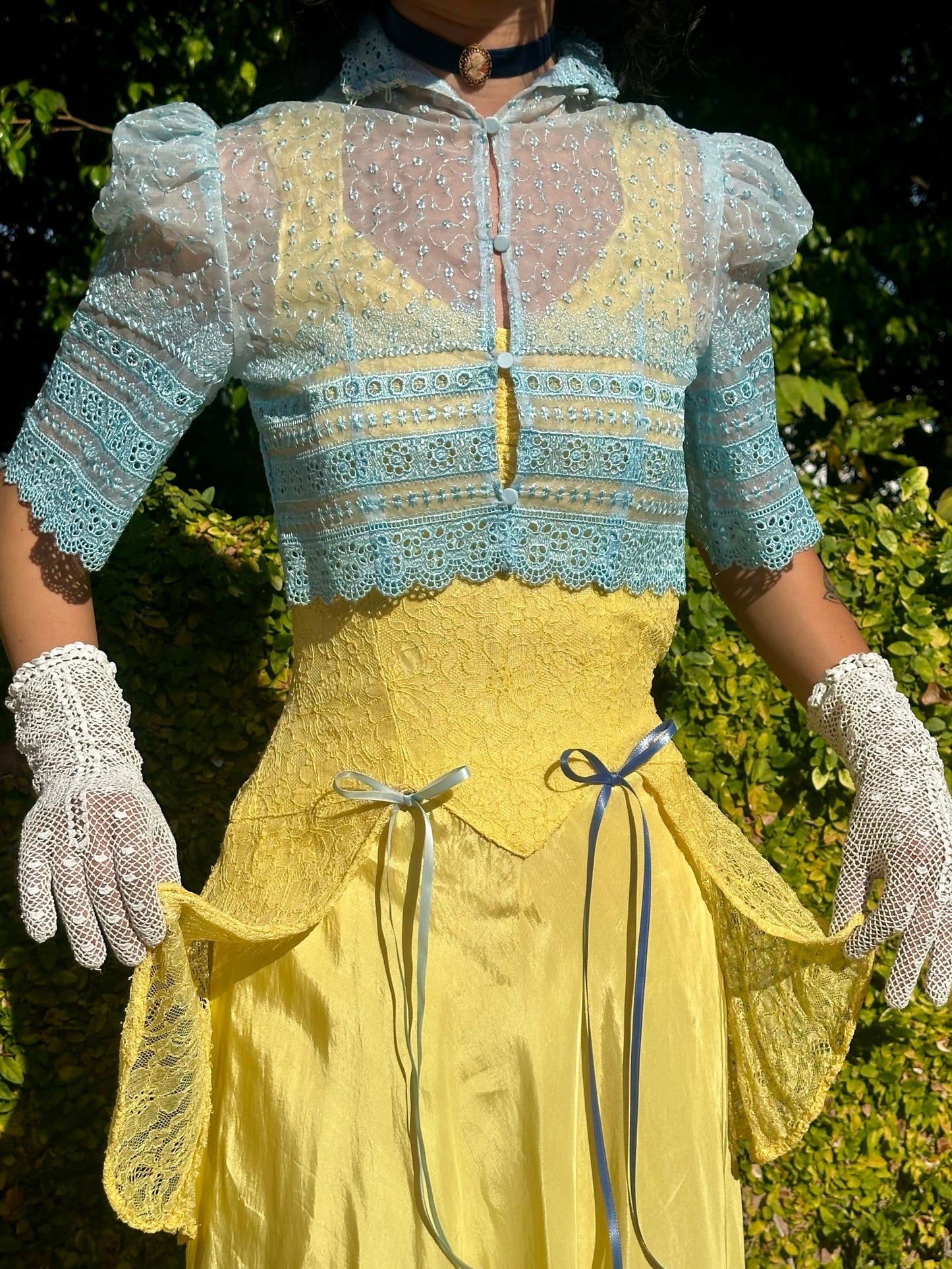 Person wearing a vintage-style yellow dress with a blue lace top against a green outdoor background.