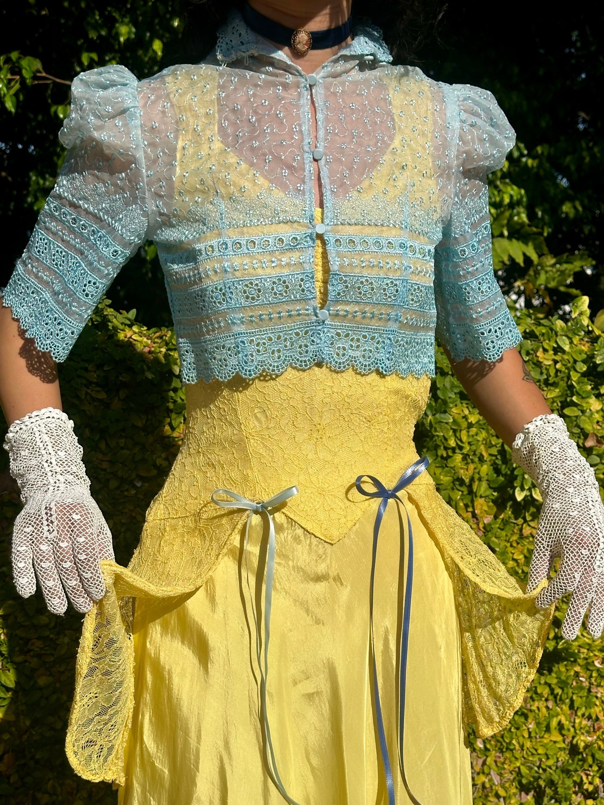 Person wearing a vintage-style yellow dress with a blue lace top against a green outdoor background.