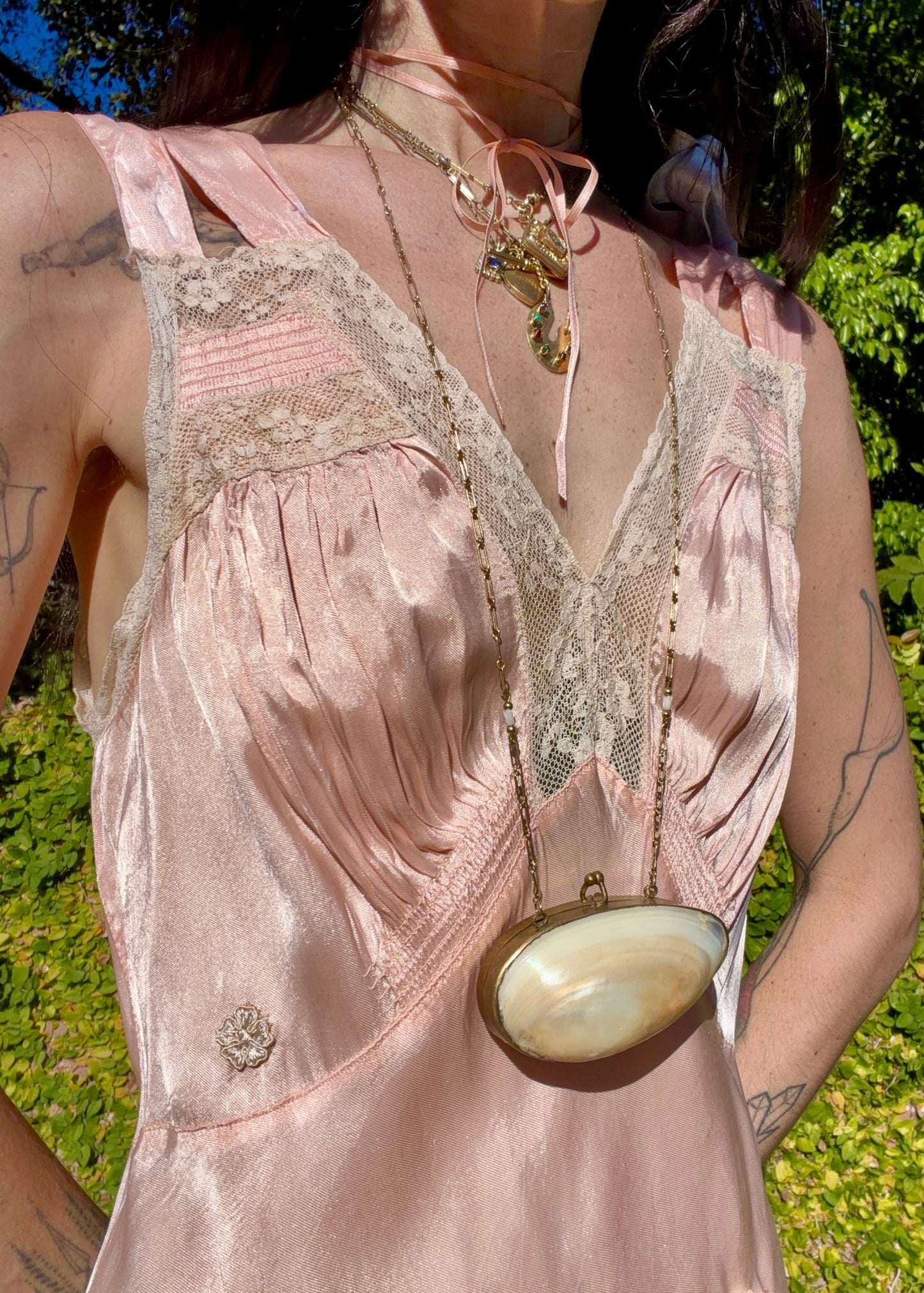 A woman wearing an antique 1930's baby pink silk slip floor-length pale blush satin gown with sheer panels and small floral appliqués, standing outdoors in front of a green hedge and stone path.