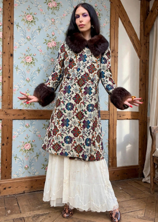 A woman is wearing a vintage 1960's / 1970's tapestry coat with a large dark brown faux fur collar and matching faux fur cuffs, posed in front of a floral wallpapered wall.