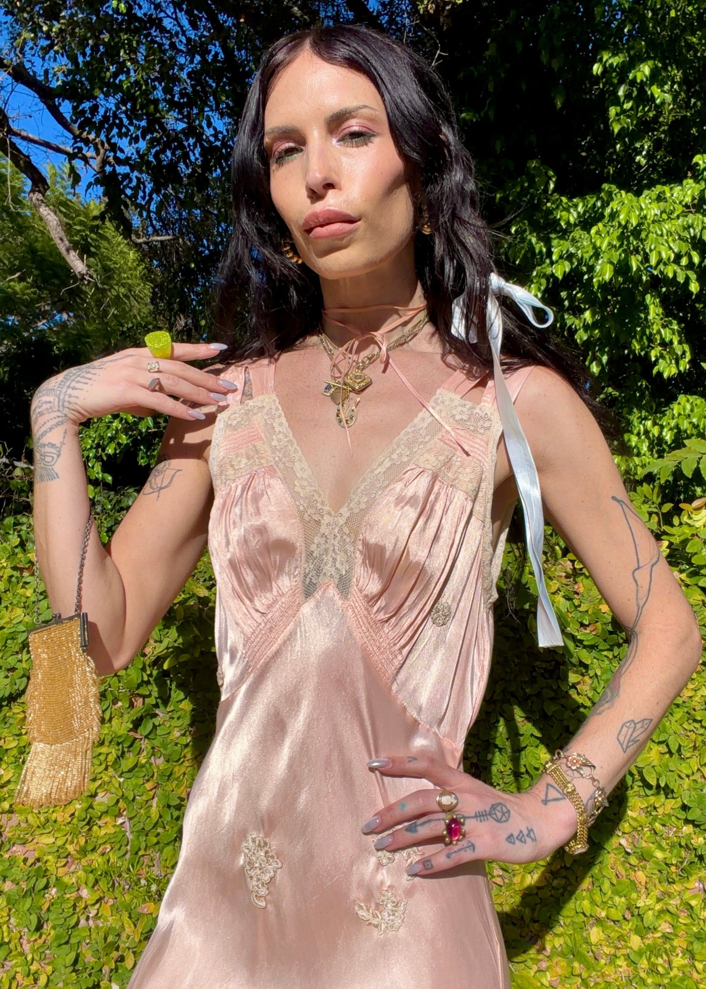 A woman wearing an antique 1930's baby pink silk slip floor-length pale blush satin gown with sheer panels and small floral appliqués, standing outdoors in front of a green hedge and stone path.
