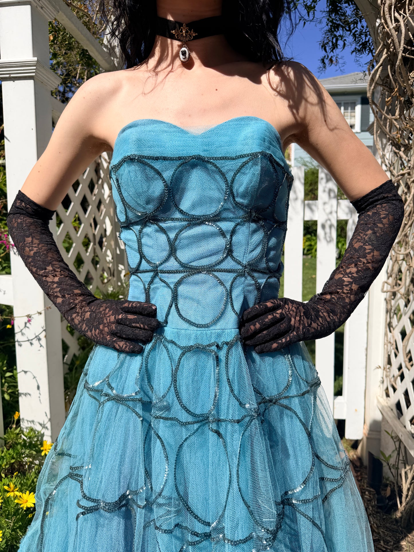Woman in a 1950's blue strapless ball gown with black gloves standing in a garden.