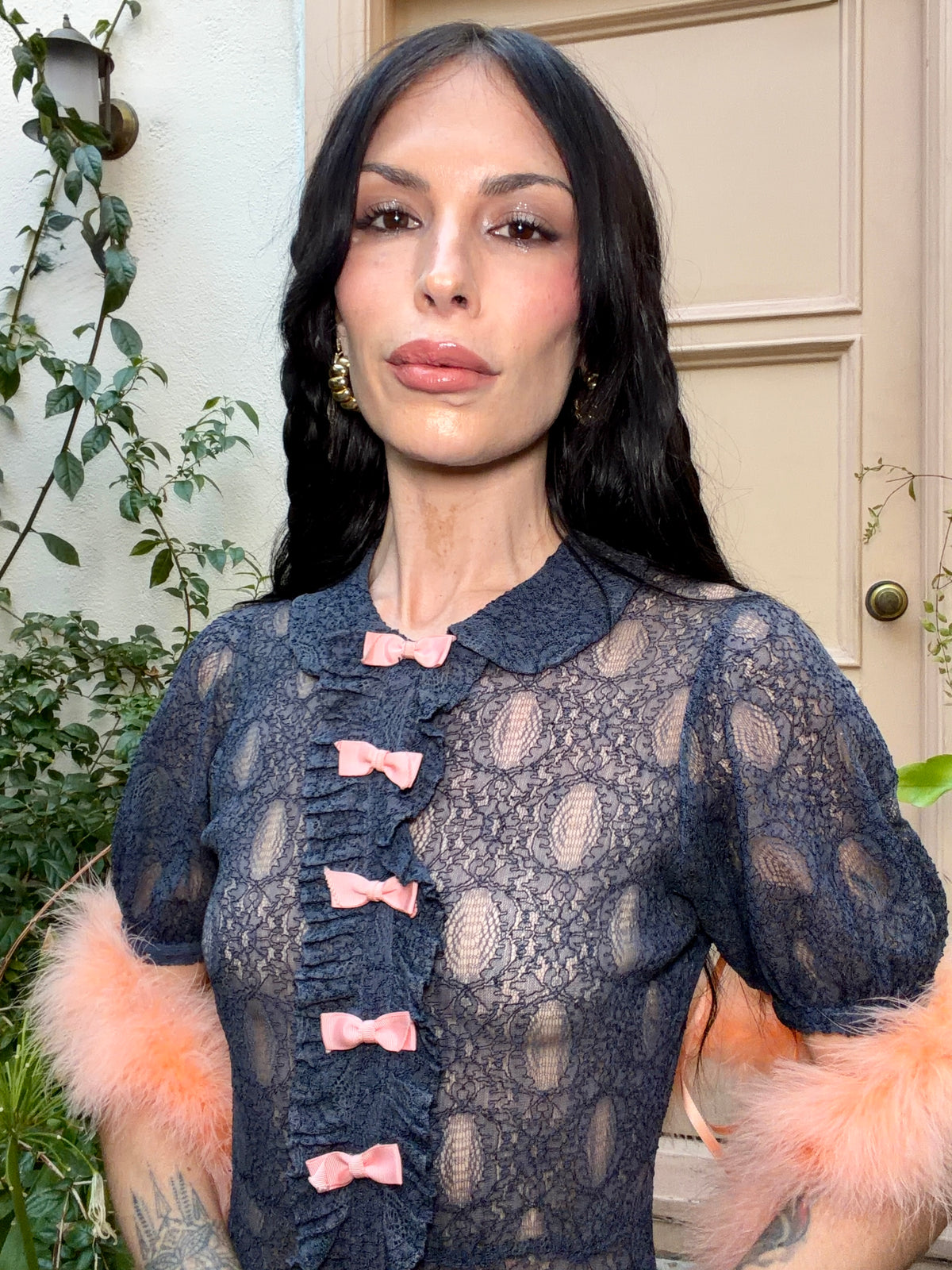 A woman wearing an antique 1930's navy blue lace dress with baby pink bows placed from the neckline all the way to the bottom of the dress, right in the center of the dress. Romantic puff sleeves, bias cut silhouette. 