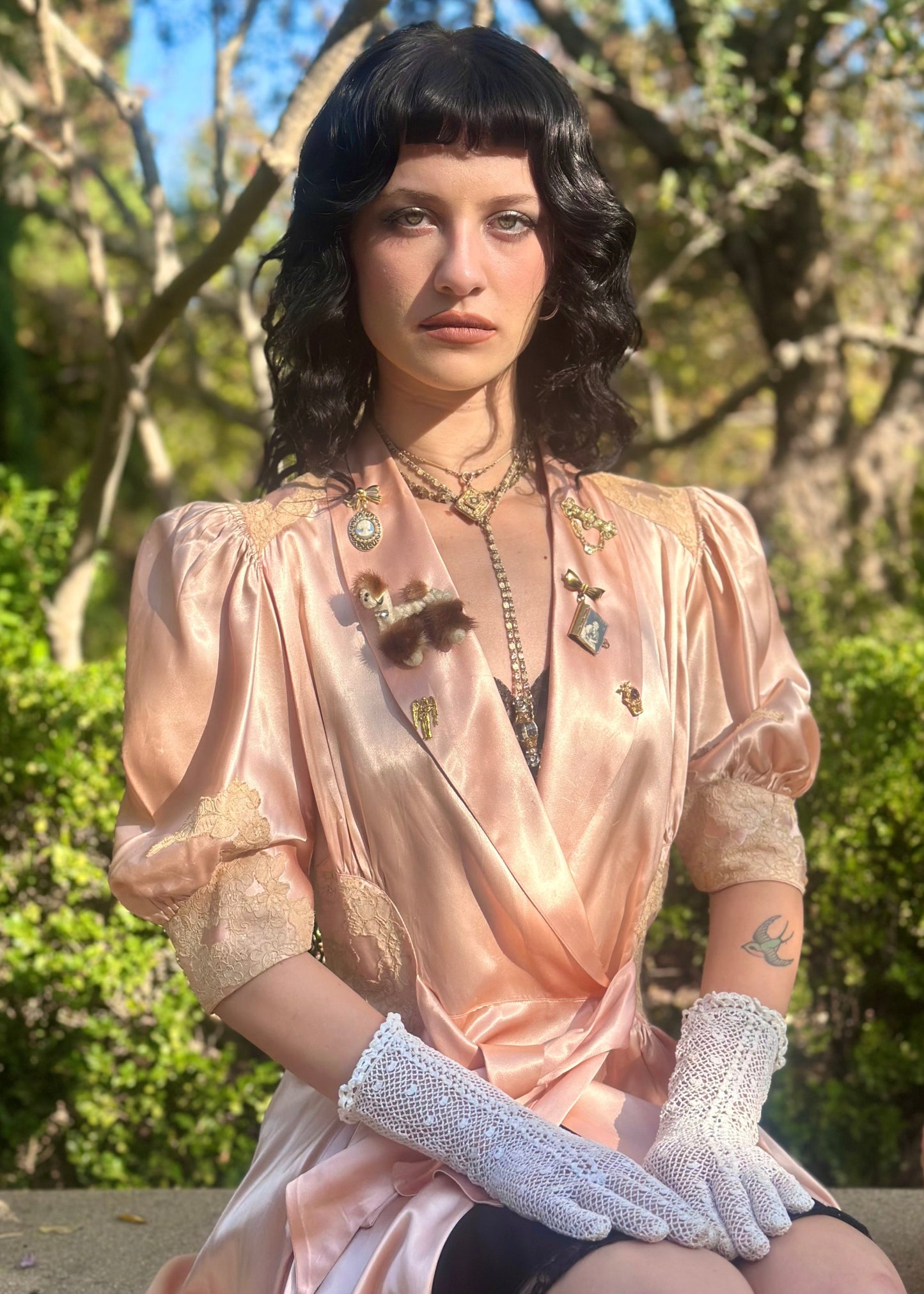 Woman in a 1940's dressing gown in peach pink with antique brooches standing between two metal arches in a garden.