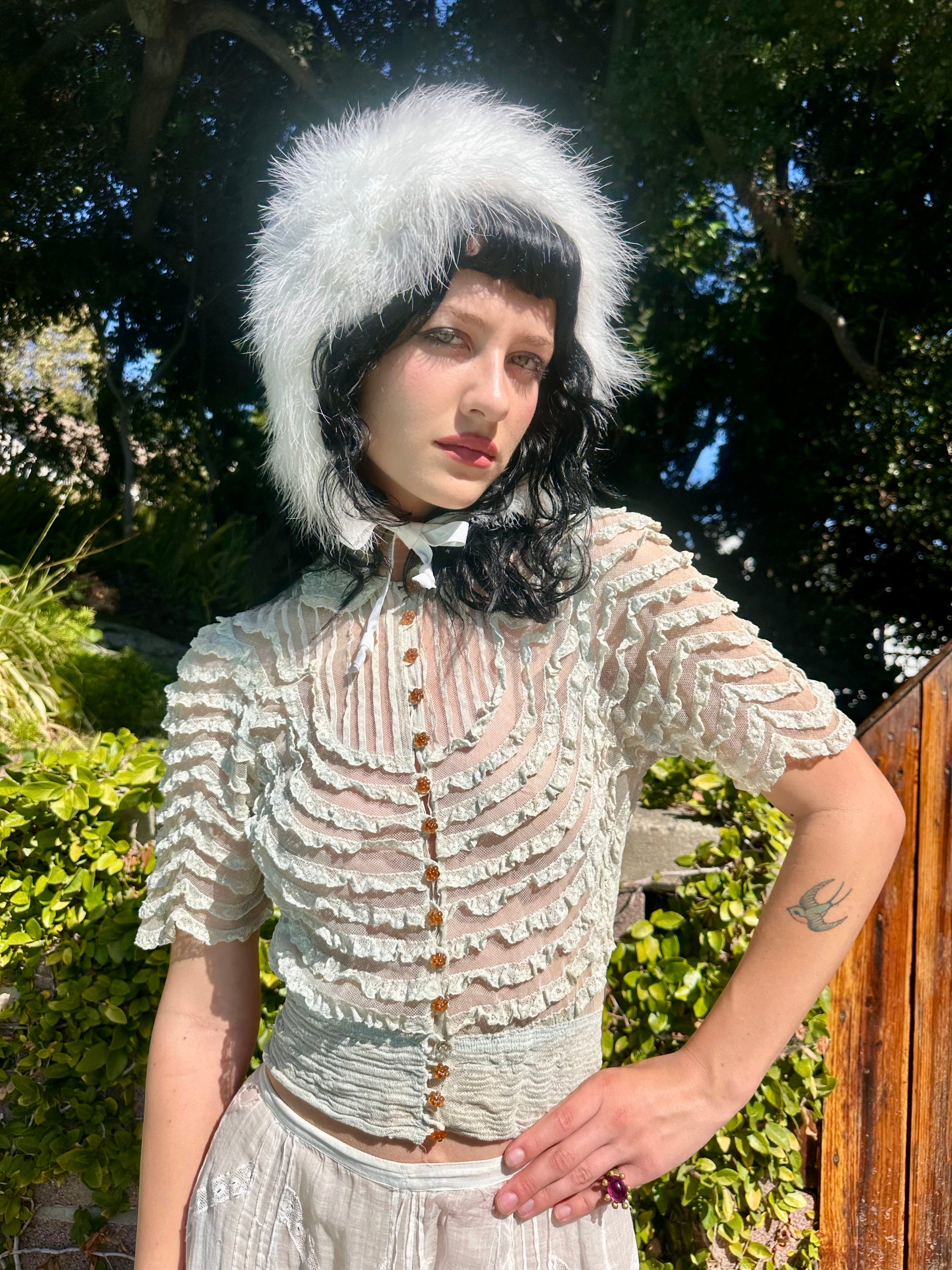 Person wearing a white lace top and fur hat outdoors with greenery in the background
