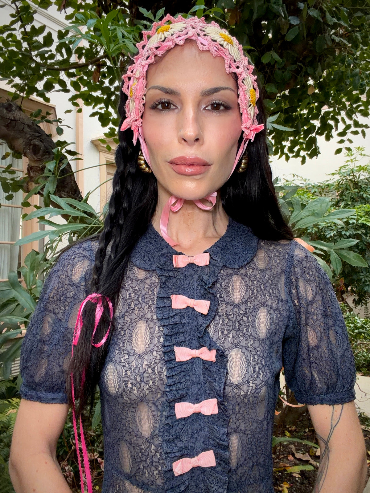 A woman wearing an antique 1930's navy blue lace dress with baby pink bows placed from the neckline all the way to the bottom of the dress, right in the center of the dress. Romantic puff sleeves, bias cut silhouette. 