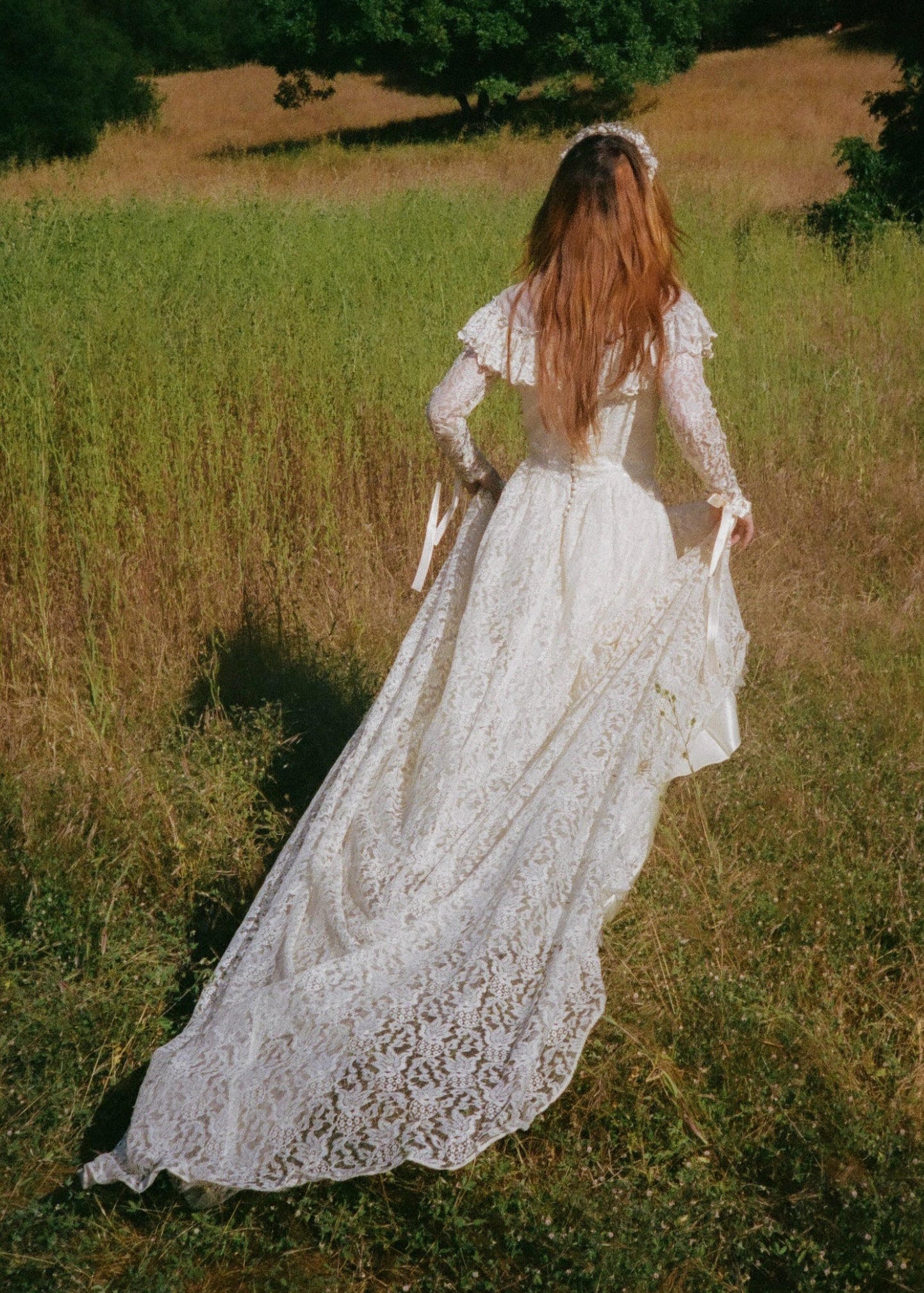 Woman in a white lace dress walking through a field with trees in the background