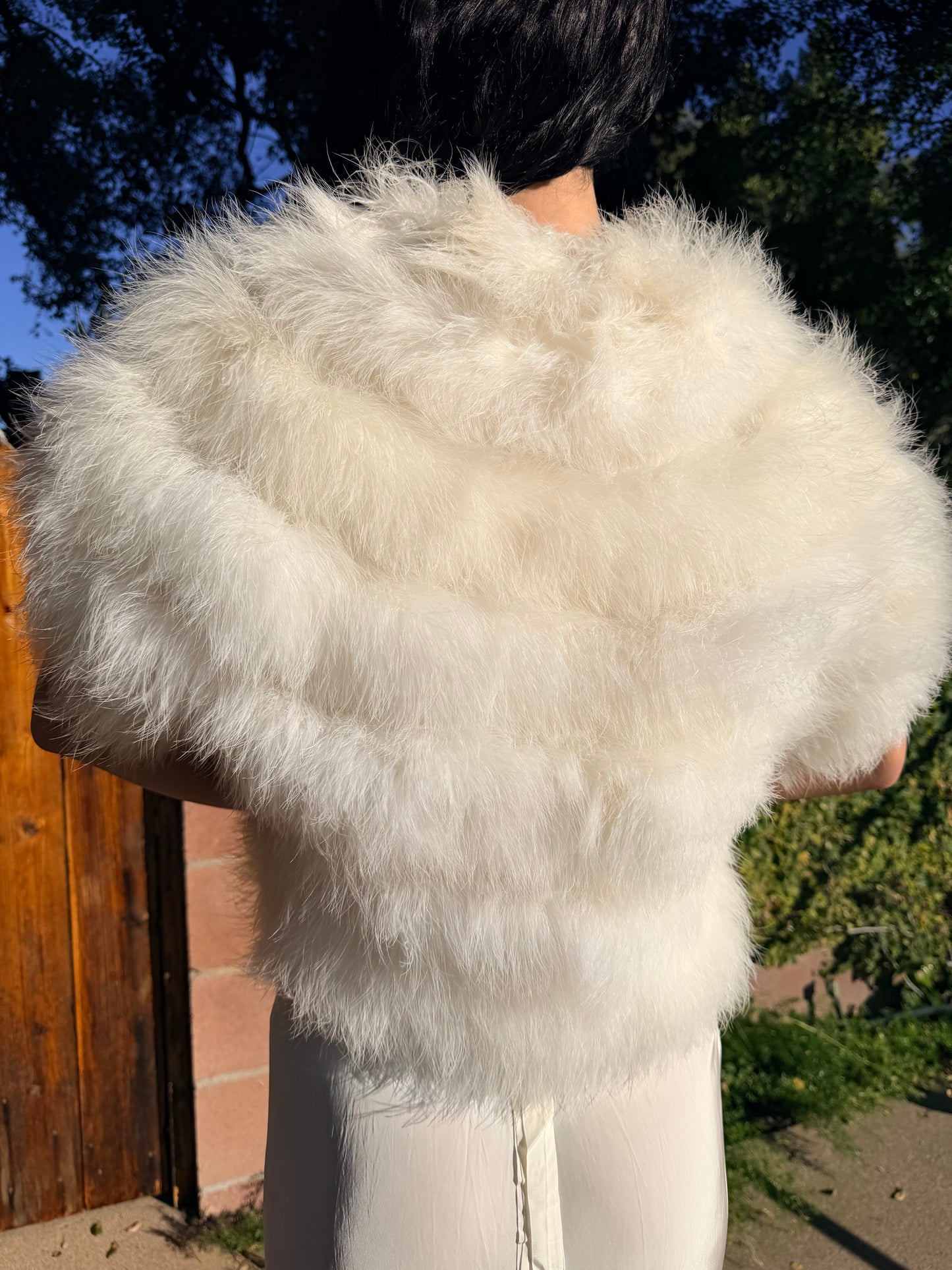 Person wearing a white marabou vest outdoors with trees and a wooden fence in the background