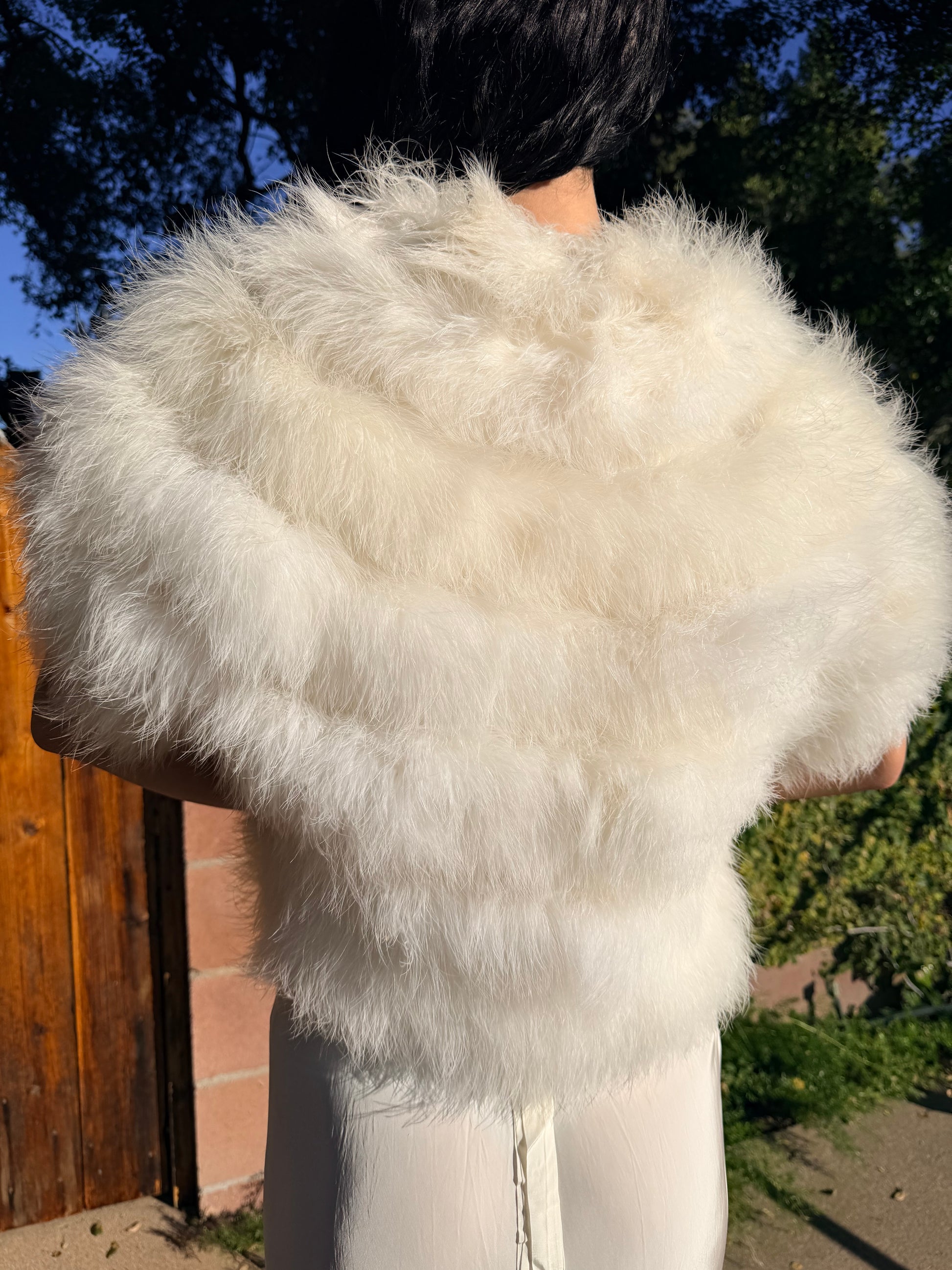 Person wearing a white marabou vest outdoors with trees and a wooden fence in the background