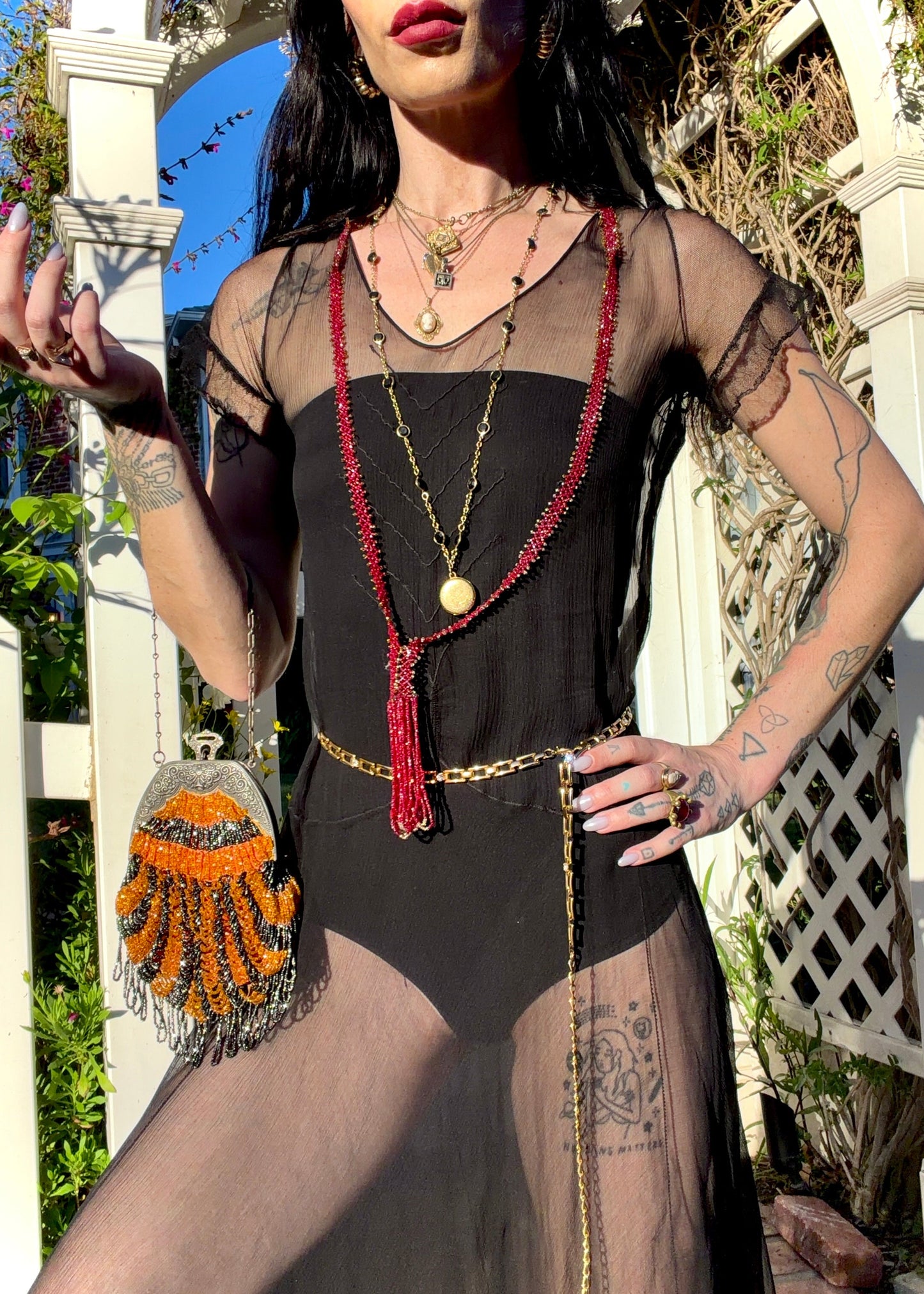 A woman stands outdoors under a white arched garden gate wearing a sheer black antique 1920's dress with a long black mesh skirt, layered gold necklaces, a gold chain belt, and beige high heels.