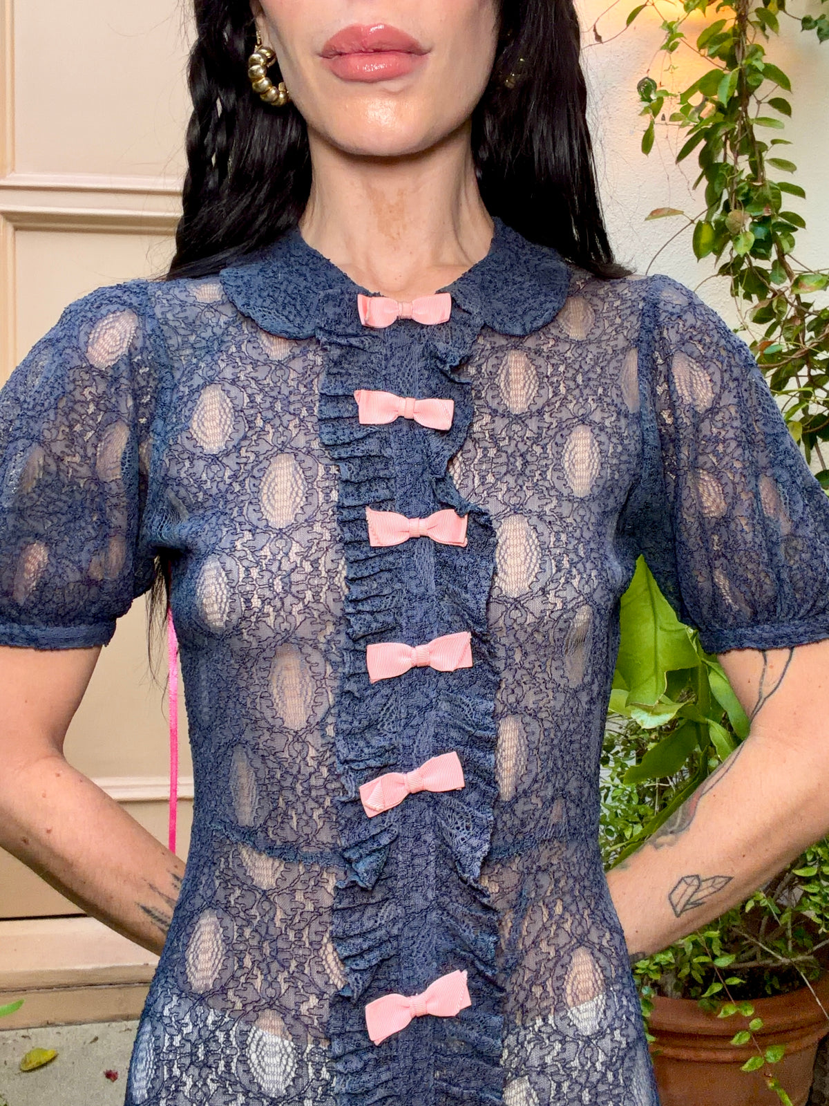 A woman wearing an antique 1930's navy blue lace dress with baby pink bows placed from the neckline all the way to the bottom of the dress, right in the center of the dress. Romantic puff sleeves, bias cut silhouette. 