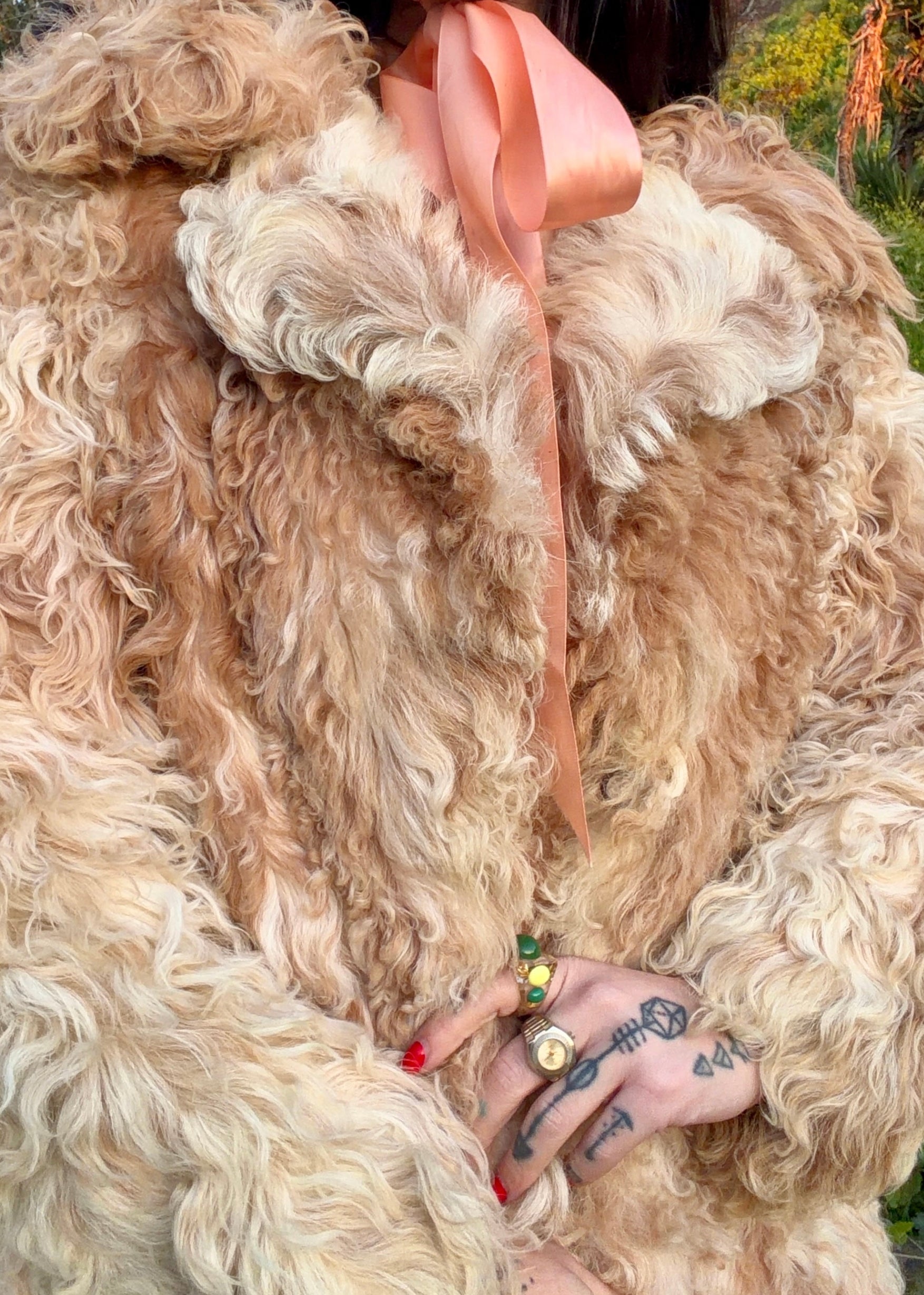 Person wearing a 1970's curly Mongolian and Tibetan sheep's fur coat in fluffy beige and brown colors in a natural setting with trees and grass.