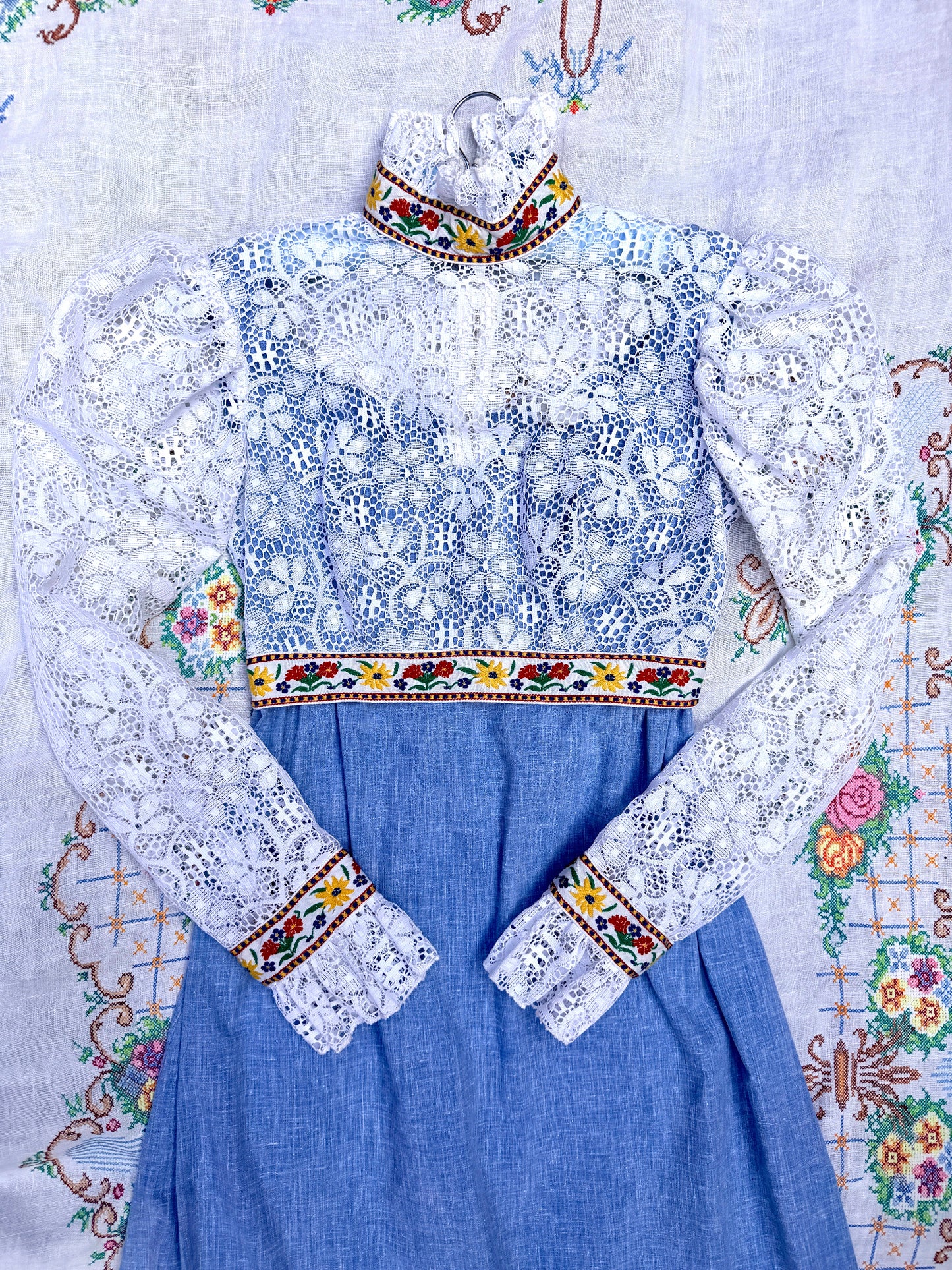 A traditional Ukrainian embroidered dress with a light blue chambray skirt, a white lace overlay bodice, and colorful embroidered borders, laid flat on a patterned white textile background.