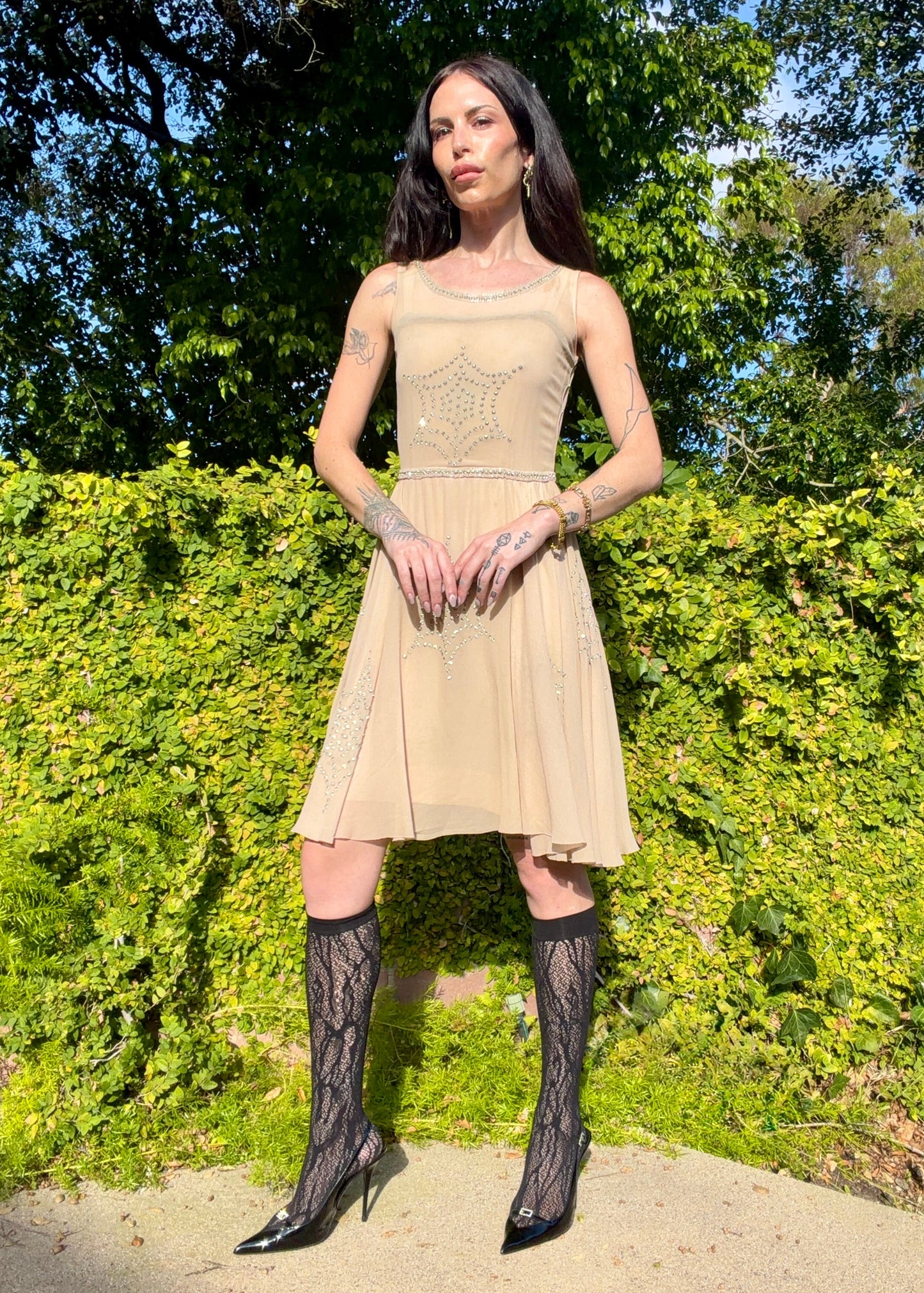Woman in a beige dress with black lace stockings standing outdoors against green foliage.