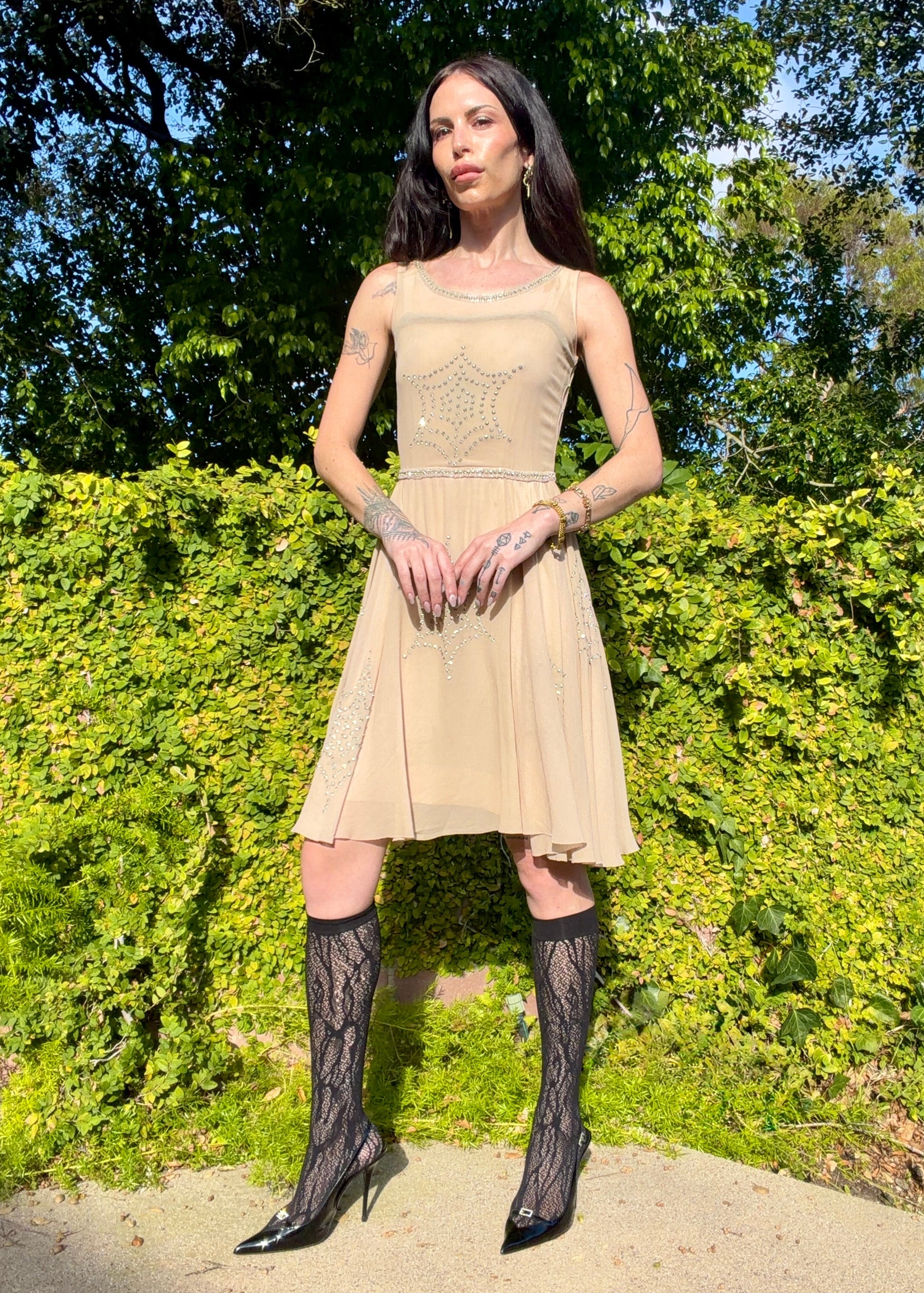 Woman in a beige dress with black lace stockings standing outdoors against green foliage.