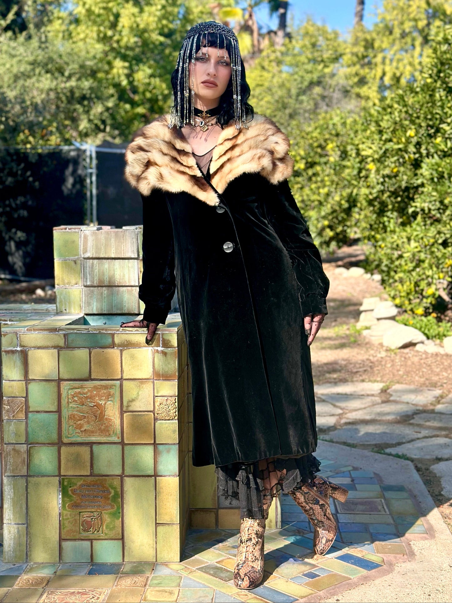 Person in an antique 1930s black coat with fur collar standing outdoors near a fountain.