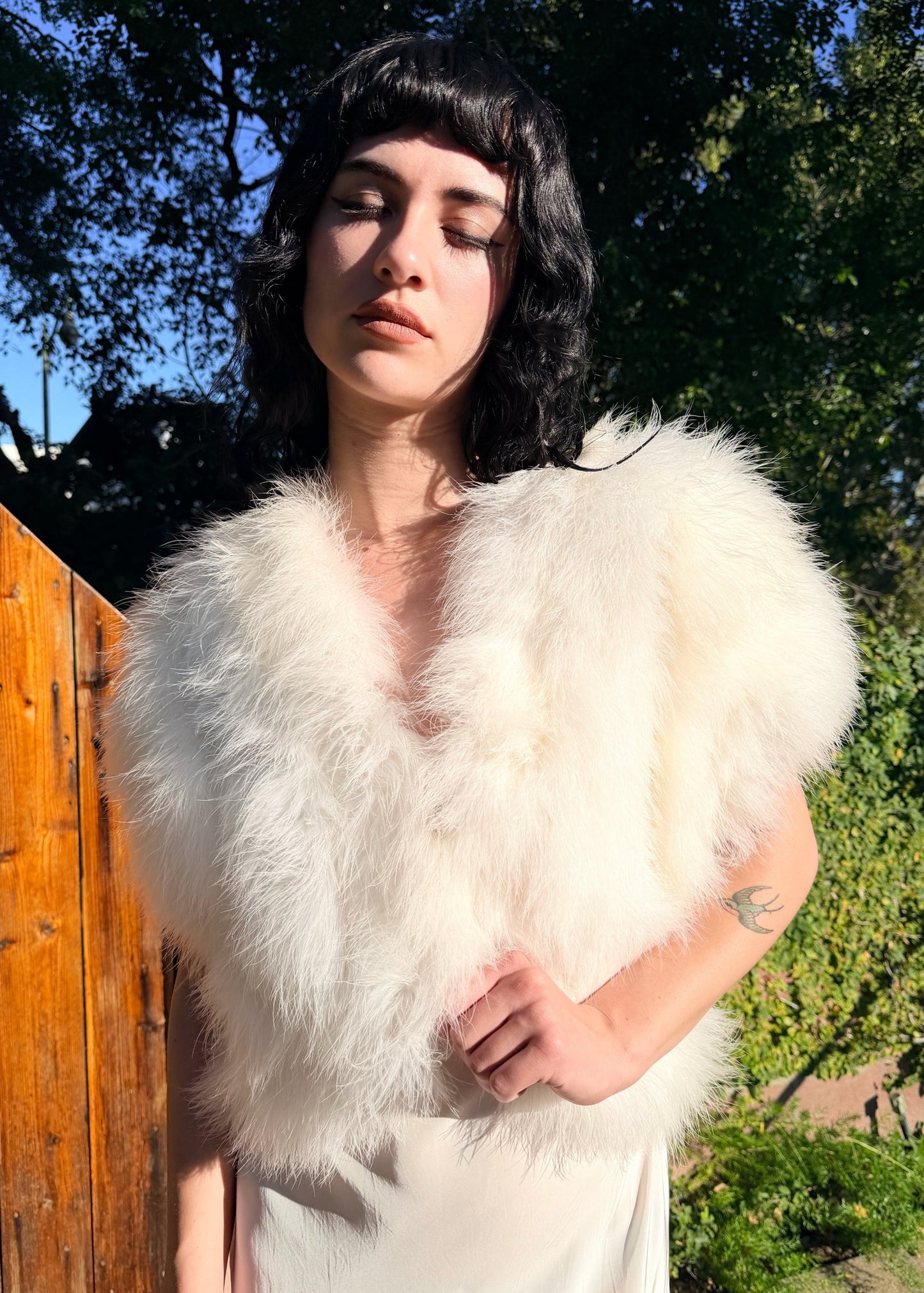 Person wearing a white marabou vest outdoors with trees and a wooden fence in the background