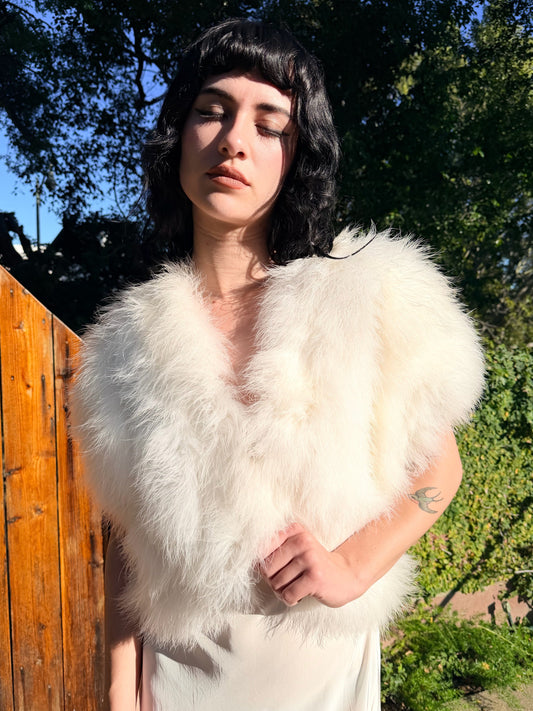 Person wearing a white marabou vest outdoors with trees and a wooden fence in the background