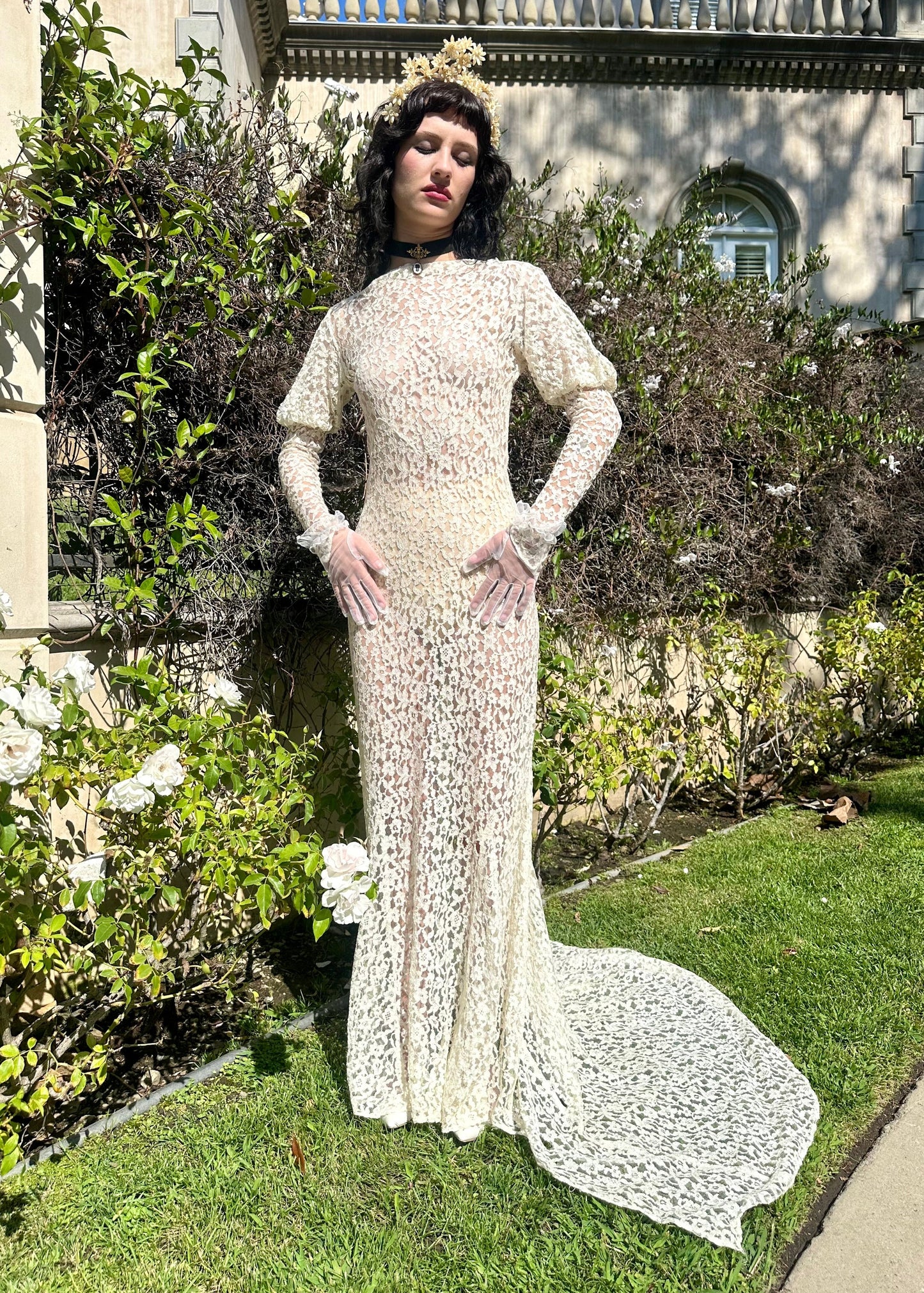 Woman in an antique 1930s white lace wedding dress standing in a garden with a classical building in the background