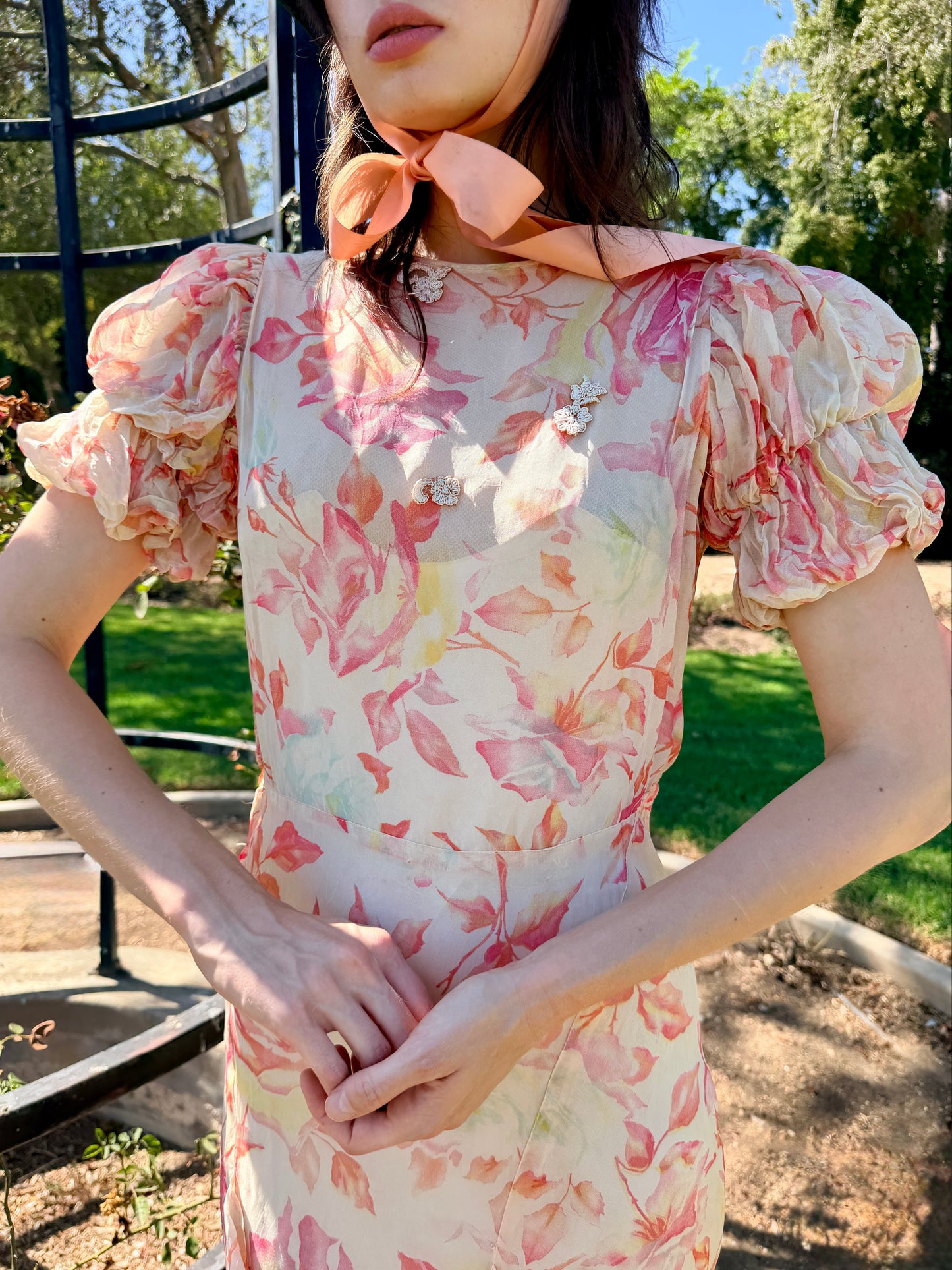 A woman wearing a cream-colored dress with a pink and orange floral print, featuring puff sleeves and a matching pink neck scarf, standing outdoors in a garden setting.