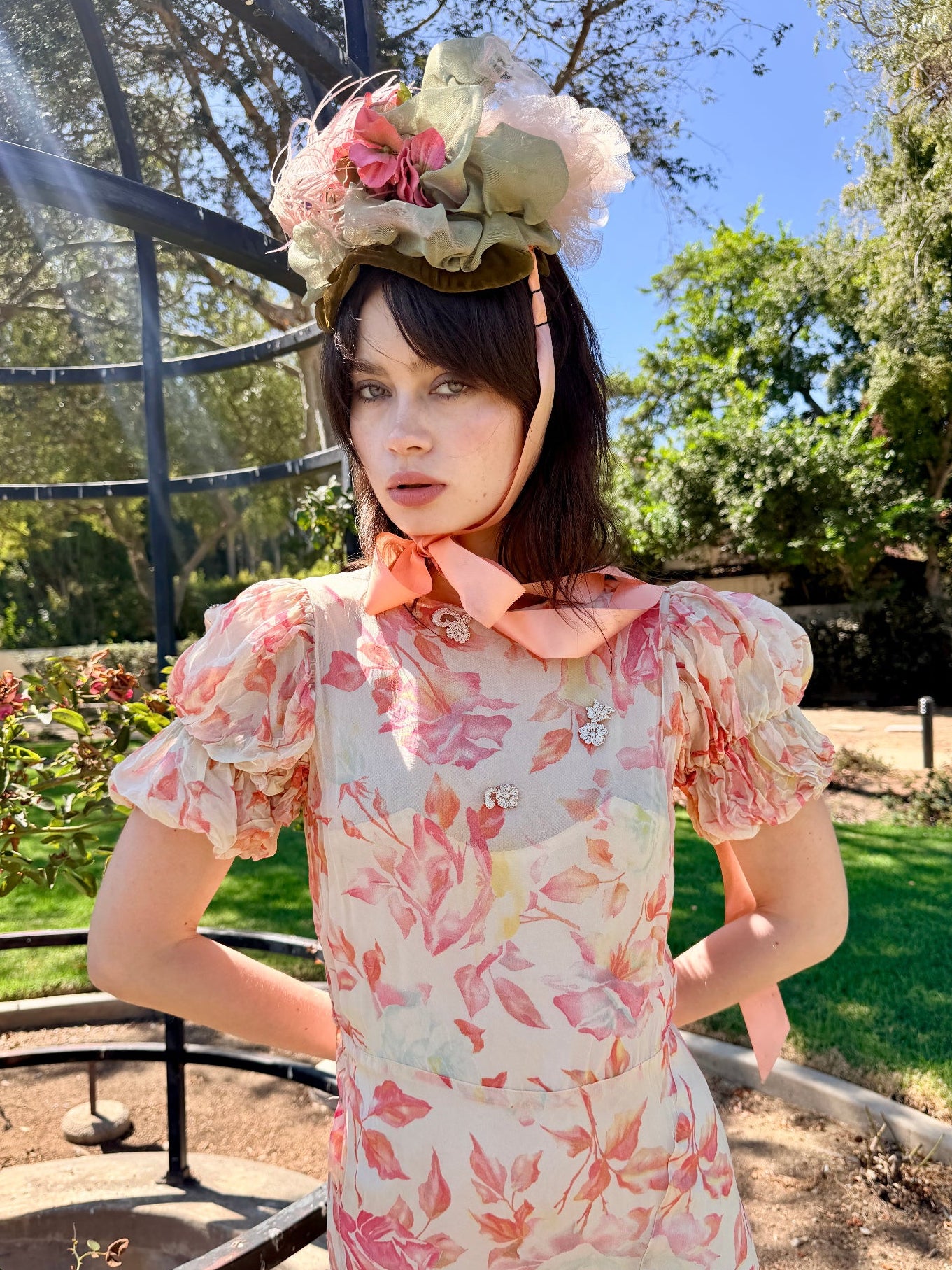 A woman wearing a floral print dress with puff sleeves and a large decorative flower headpiece, standing outdoors in a garden setting.