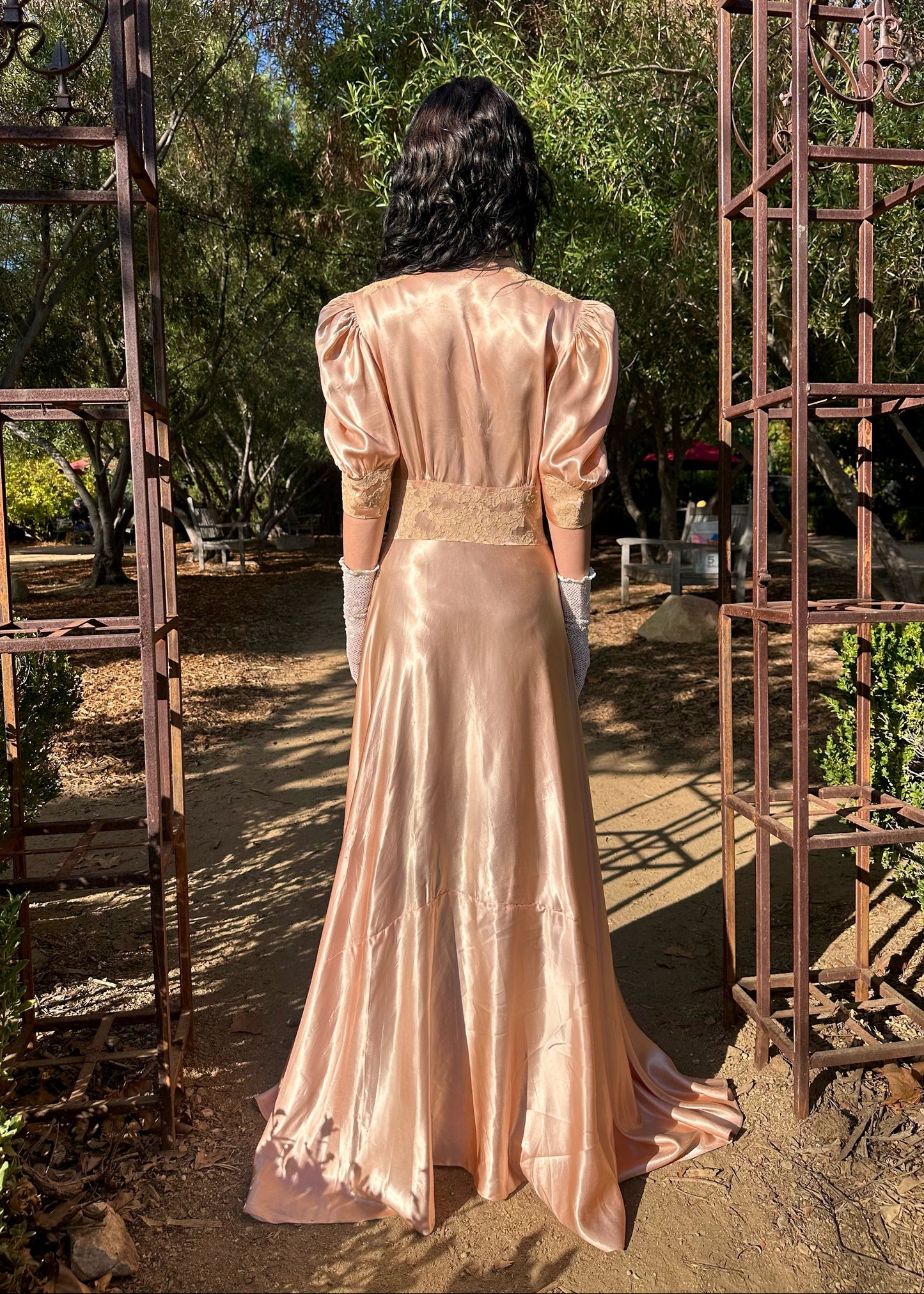 Person in a long, light pink dress standing between two metal arches in a garden setting.