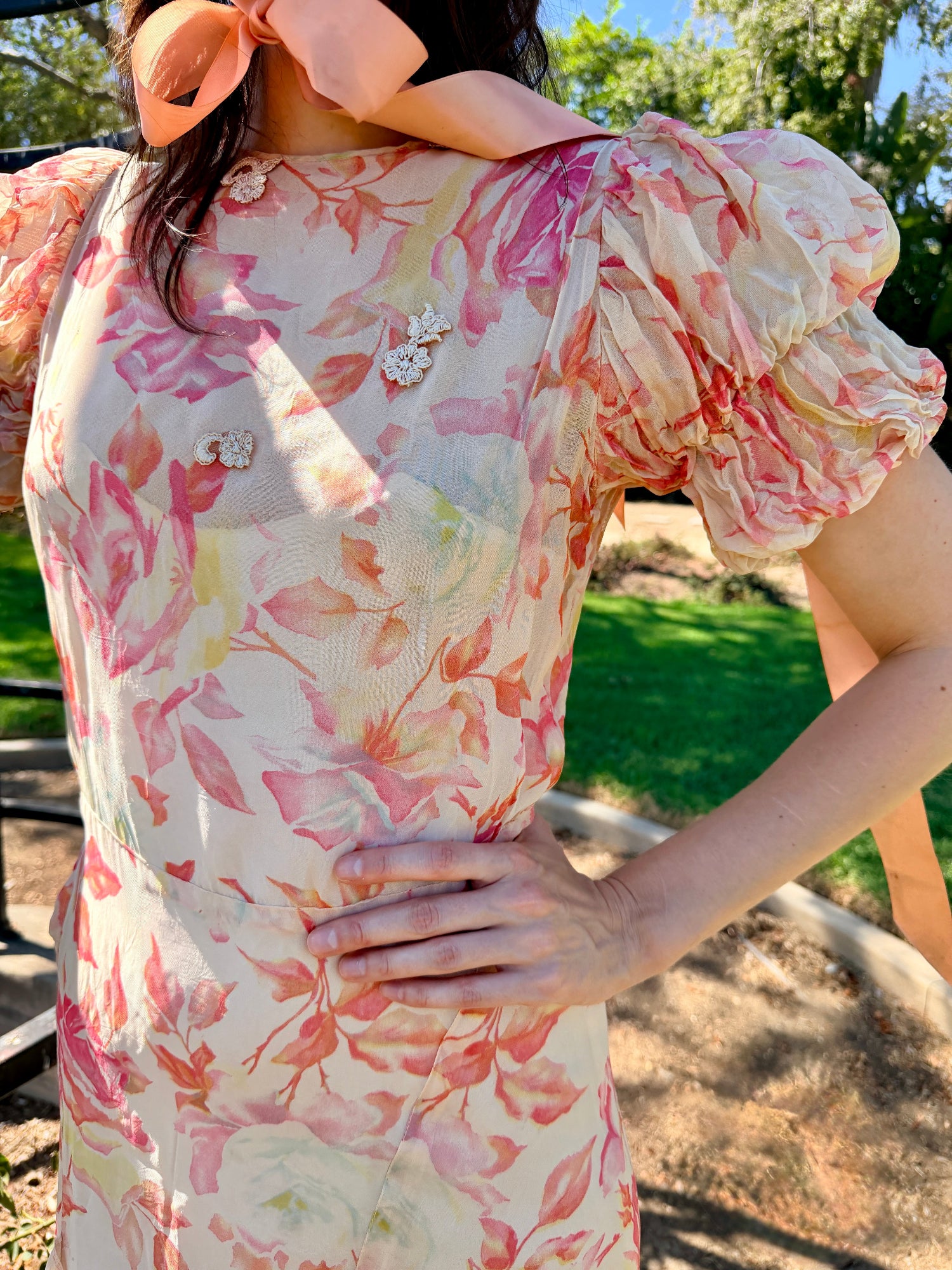 A woman wearing a short-sleeve cream-colored dress with a pink and peach floral print, featuring ruffled sleeves and decorative floral embellishments on the bodice, photographed outdoors in a garden setting.