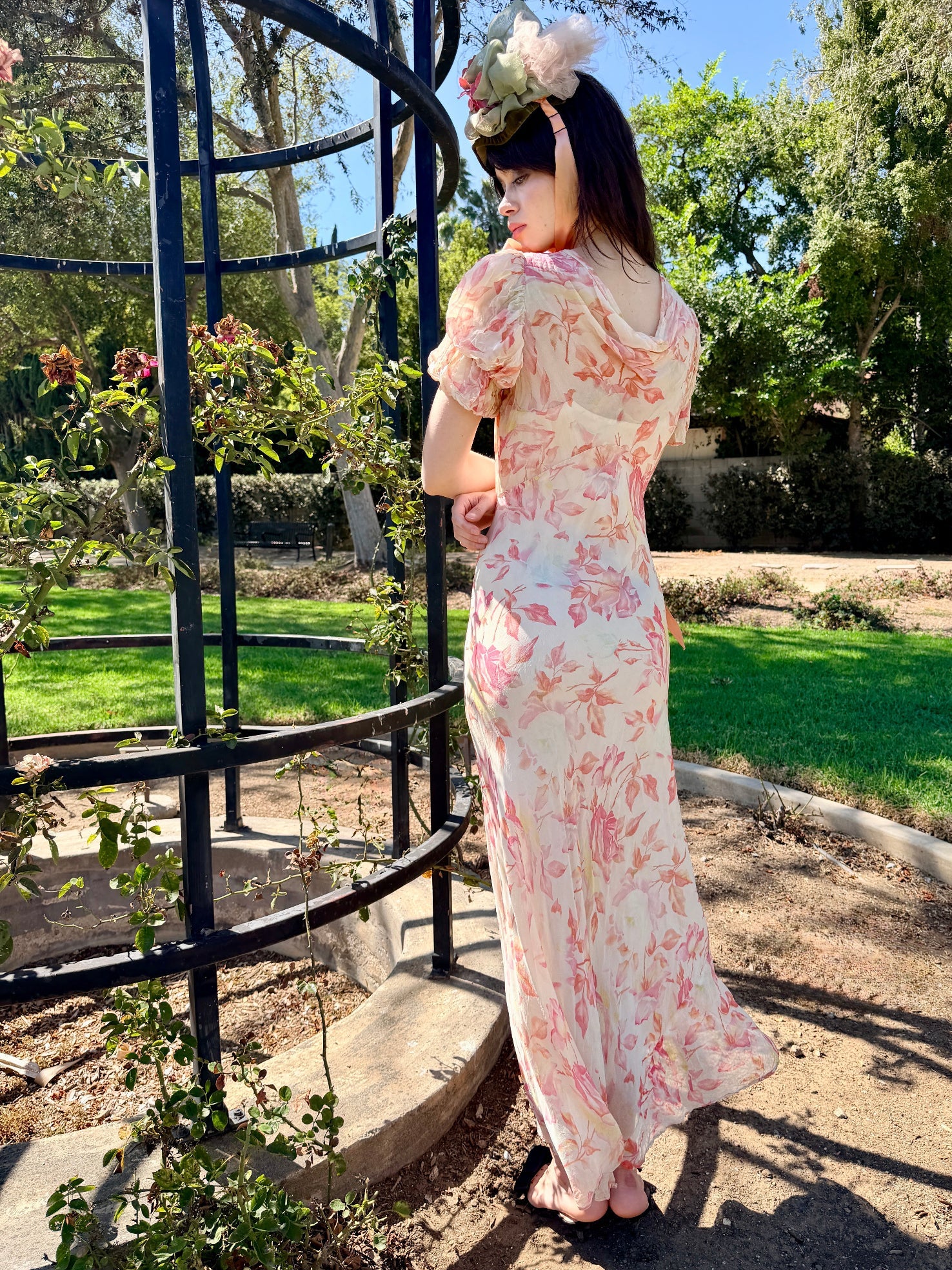 A woman stands outdoors in a garden next to a black metal gazebo, wearing a light pink and white floral maxi dress with puff sleeves and a matching floral headband.