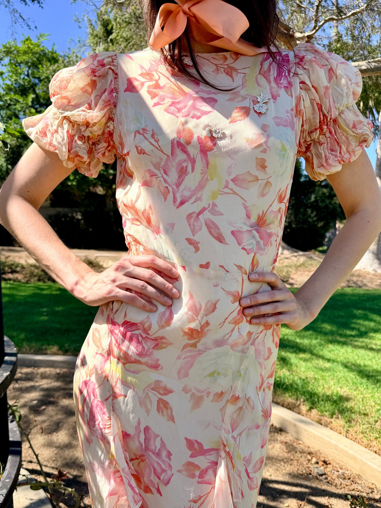A woman wearing a short-sleeve cream dress with a pink and peach floral print, standing outdoors in a garden with her hands on her hips.