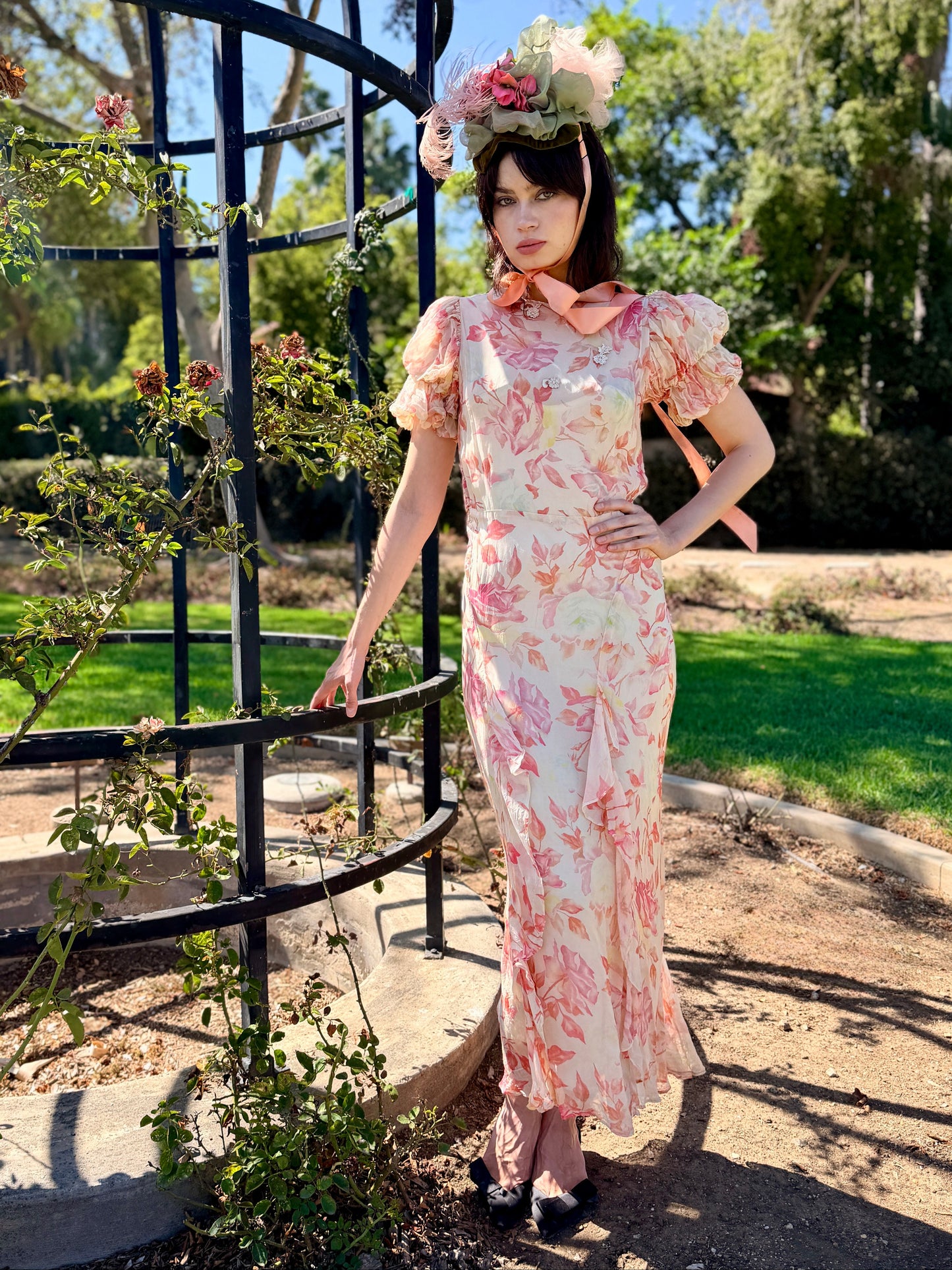 A woman stands outdoors in a garden next to a black metal trellis, wearing a long cream dress with a pink floral print, a pink bow at the collar, and a large pink and white flower crown.