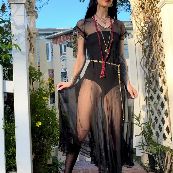 A woman stands outdoors under a white arched garden gate wearing a sheer black antique 1920's dress with a long black mesh skirt, layered gold necklaces, a gold chain belt, and beige high heels.