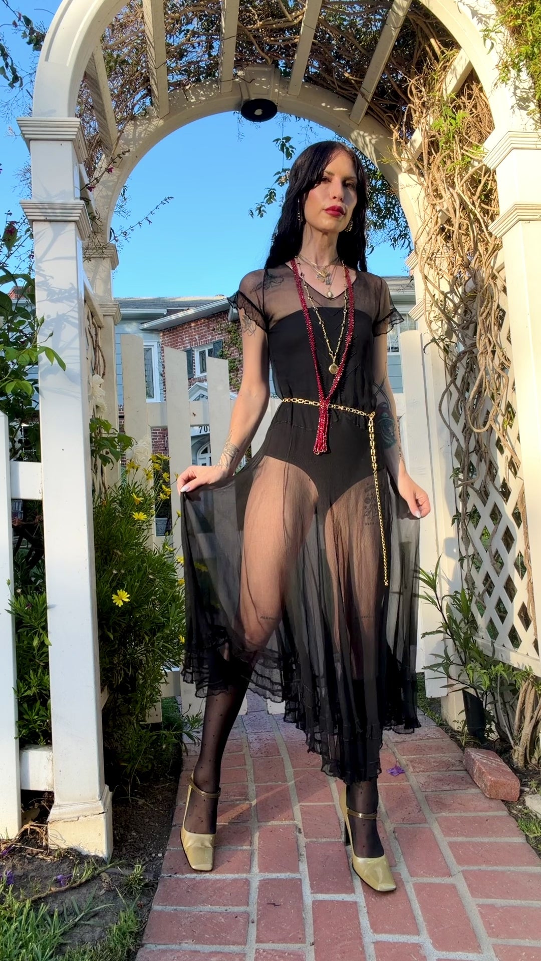 A woman stands outdoors under a white arched garden gate wearing a sheer black antique 1920's dress with a long black mesh skirt, layered gold necklaces, a gold chain belt, and beige high heels.