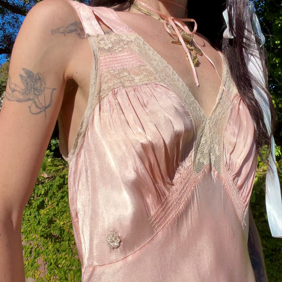 A woman wearing an antique 1930's baby pink silk slip floor-length pale blush satin gown with sheer panels and small floral appliqués, standing outdoors in front of a green hedge and stone path.