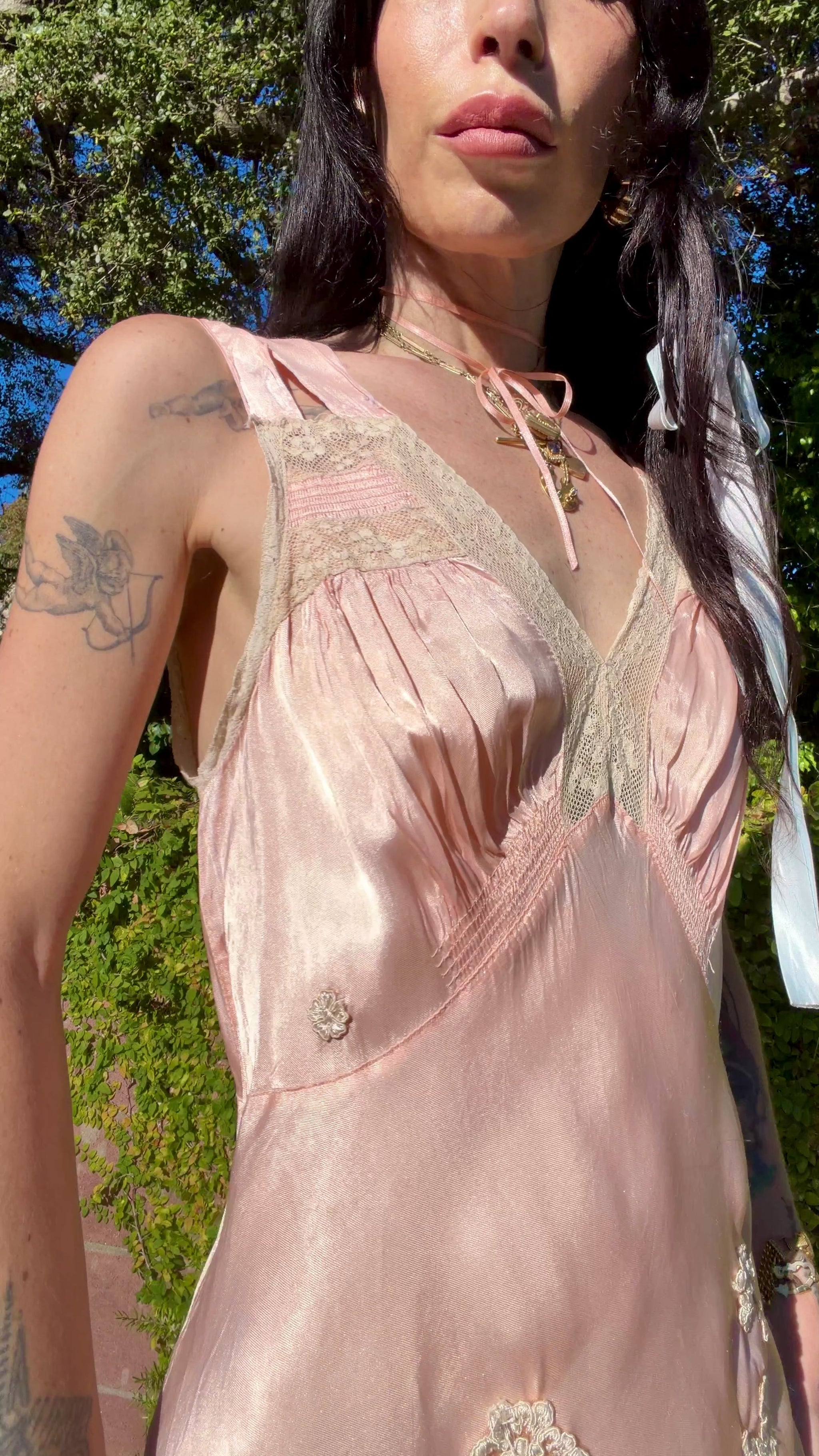 A woman wearing an antique 1930's baby pink silk slip floor-length pale blush satin gown with sheer panels and small floral appliqués, standing outdoors in front of a green hedge and stone path.