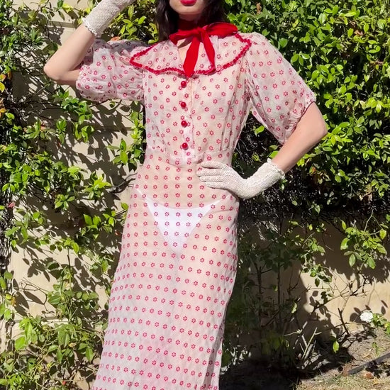 A woman wearing an 1930's silk organdy  and white and red flower maxi dress with red trim, white gloves, a black hat, and red shoes stands outdoors in front of a stone building and green hedges.