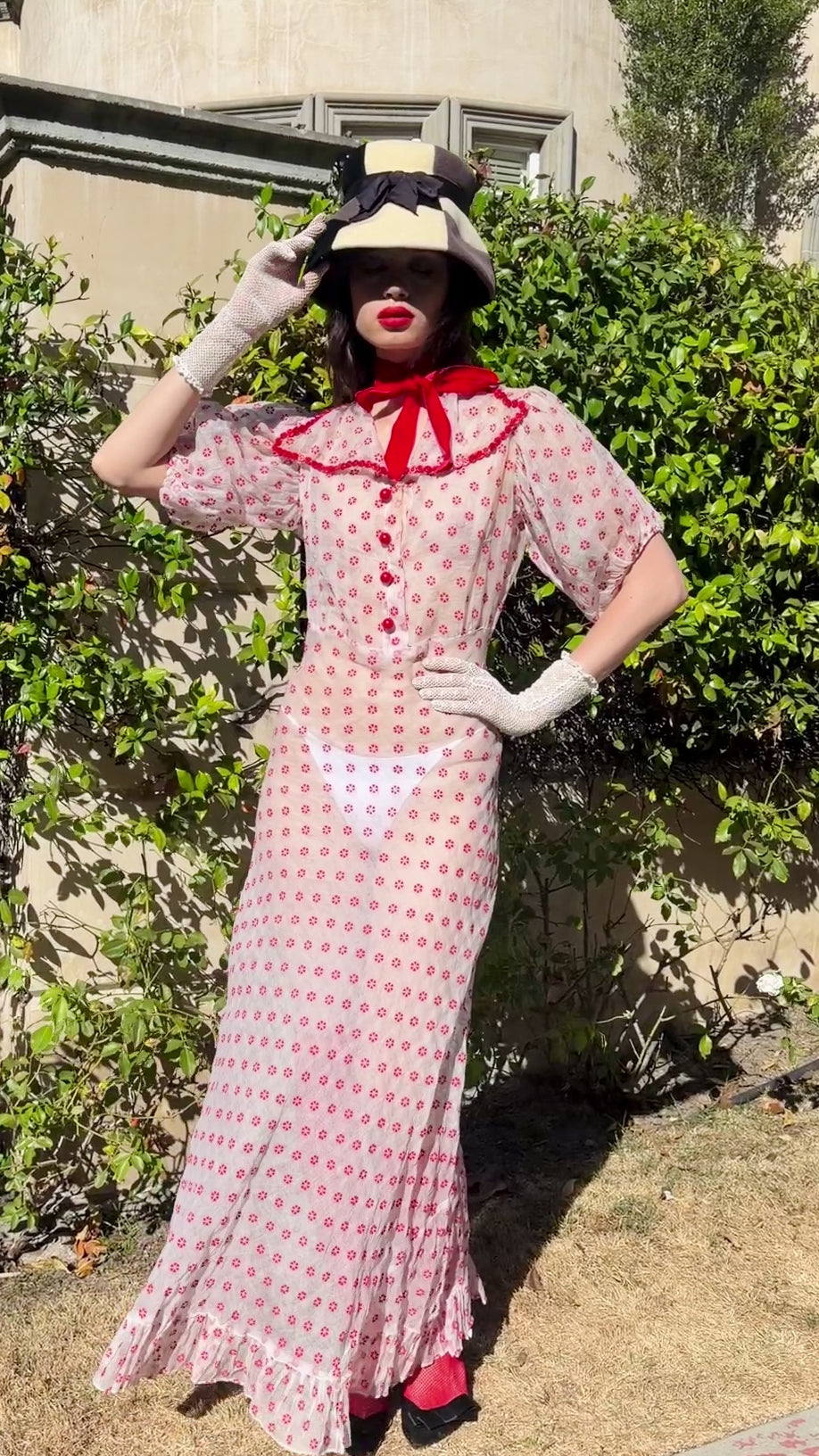 A woman wearing an 1930's silk organdy  and white and red flower maxi dress with red trim, white gloves, a black hat, and red shoes stands outdoors in front of a stone building and green hedges.