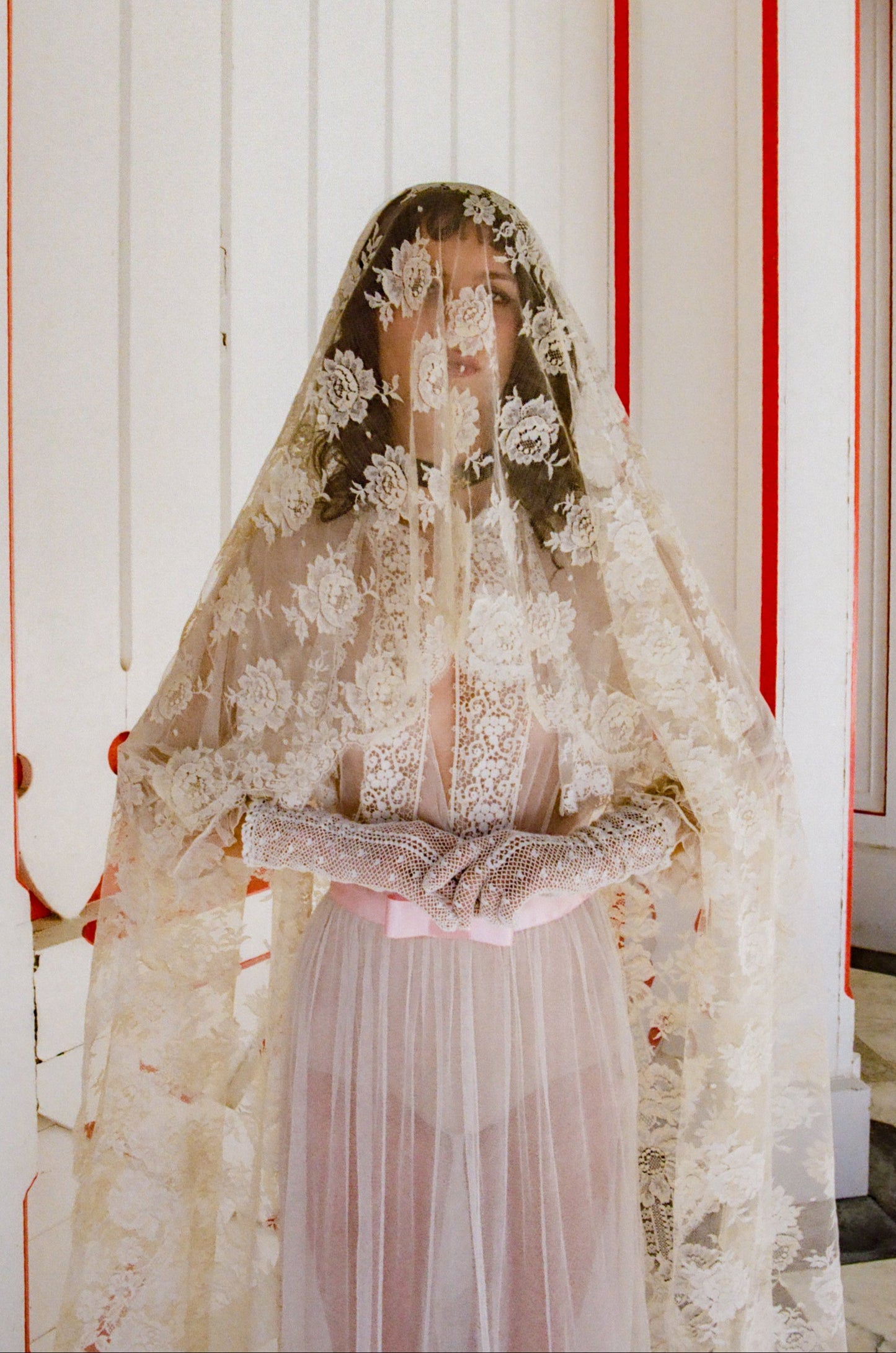 Person wearing a lace veil in an indoor setting with red and white walls.